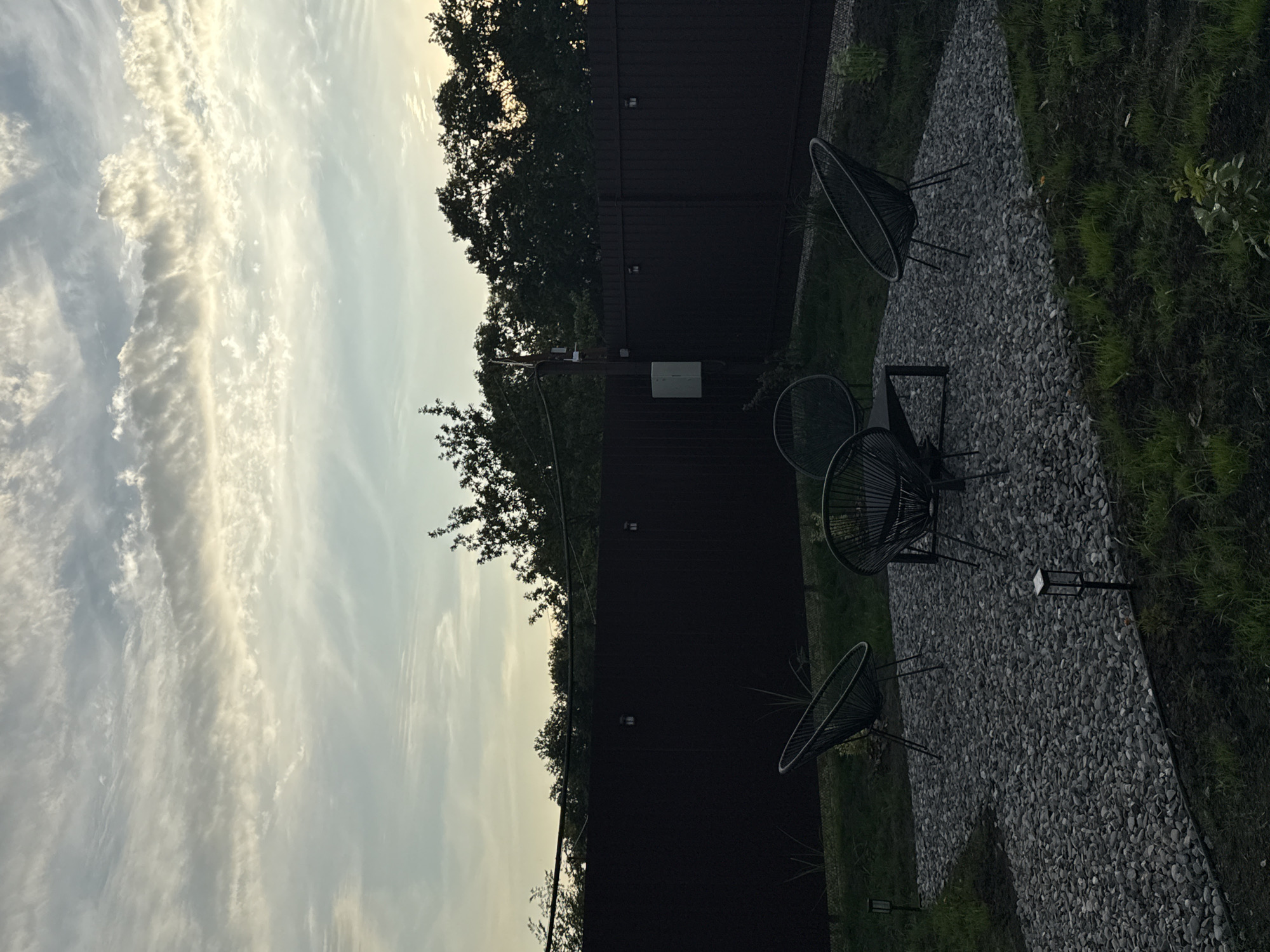 This is a vertical, outdoor shot of a backyard area at dusk. The scene features a dark, slatted fence running horizontally across the mid-ground. To the left of the fence, the sky is filled with wispy, elongated clouds illuminated by the setting sun, creating a dramatic, streaky effect. Trees with dark foliage line the top of the fence, silhouetted against the sky.
In the foreground, a pathway of dark grey gravel leads diagonally from the bottom right to the top left of the frame. On this path, there are two black, wire-frame chairs with rounded backs and a small, low black table between them. Two small, rectangular garden lights are visible along the edge of the gravel path. To the right of the gravel path, a strip of dark green grass is visible, with some lighter green foliage growing in it. Another garden light is planted in the grass near the bottom edge of the frame.
The overall mood is tranquil and slightly melancholic, fitting for the twilight hour. The light filtering through the clouds suggests a cool evening. The setting appears to be a private residence. There is no visible text in the image.
