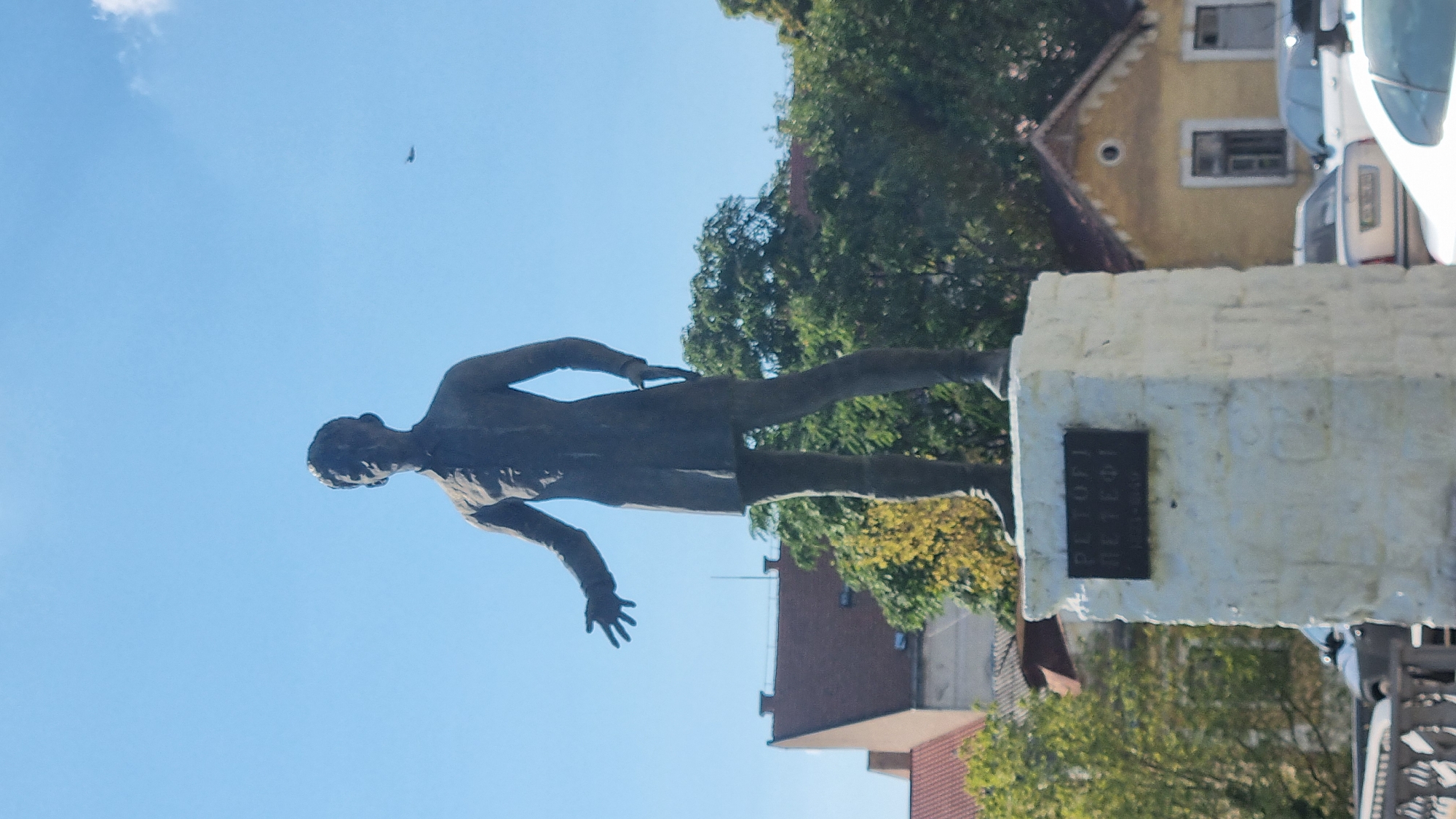 A bronze statue of a man stands on a white stone pedestal. The man is depicted in a dark, knee-length coat and trousers, with his left hand outstretched. A plaque with Cyrillic inscription is affixed to the front of the pedestal. The statue is located in a public space with buildings and trees visible in the background. The sky is clear and blue, with a small airplane in the distance. The statue is believed to be of the Hungarian poet Sándor Petőfi, and is located in Berehove, Ukraine.