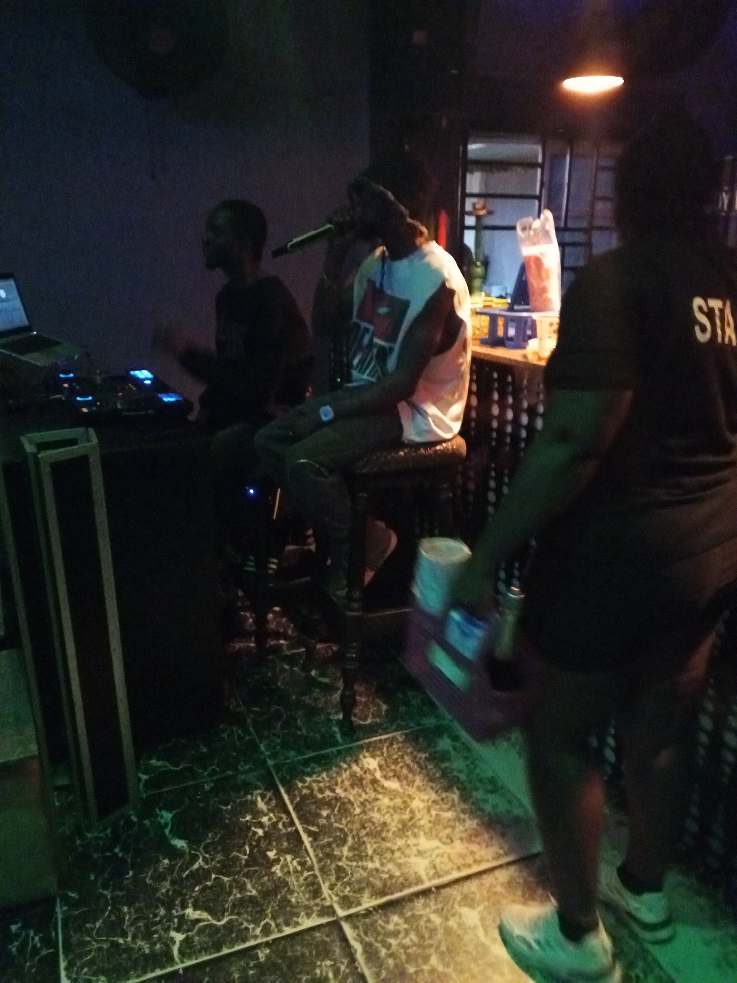 The image shows a dimly lit indoor scene, possibly a nightclub or bar in Lokoja, Nigeria. On the left, a person is operating DJ equipment on a table, with a laptop visible. In the center, another person sits on a stool, holding a microphone. The person wears a white tank top and dark pants. On the right, a "STAFF" member is visible in a dark t-shirt and shorts, carrying a tray with items, possibly drinks. The floor is tiled with a dark, marble-like pattern. There is a bar area in the background.