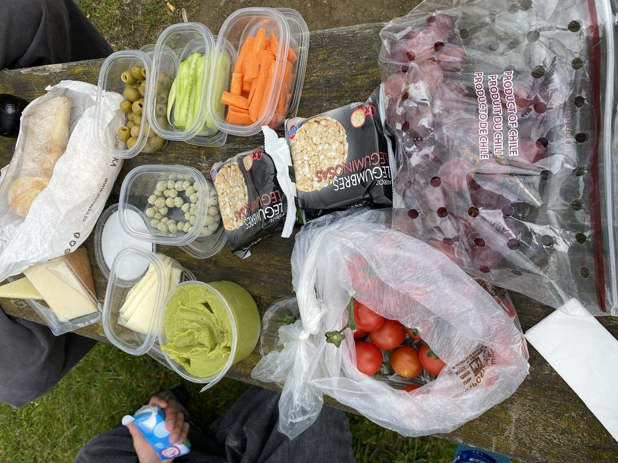 The scene shows a picnic spread on a weathered wooden surface, possibly a picnic table. The food includes a baguette, olives, sliced cucumbers and carrots, green coated spheres, rice cakes, grapes in a clear plastic bag labeled "PRODUCT OF CHILE", tomatoes in a plastic bag, sliced cheese, and green dip. A person is visible in the lower portion of the image, wearing grey pants and holding a small blue and white carton. A napkin is also visible. The food items are a mix of packaged and fresh items, suggesting a casual outdoor meal. Without other visual cues, the location in Villaviciosa, Spain is not identifiable.