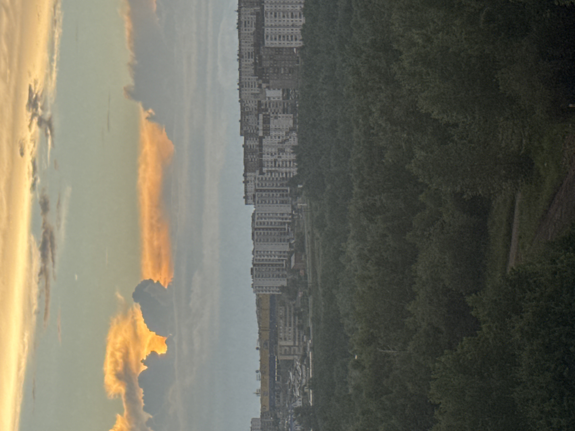 The image depicts a panoramic view of a city skyline against a dramatic sunset sky, with a dense forest in the foreground. The city of Bratsk, Russia, is identifiable by its characteristic Soviet-era panel buildings, many of which are tall apartment blocks. The skyline features a long, yellow-fronted commercial building, likely a large supermarket or retail center, in the middle ground. The sky is filled with warm hues of orange and yellow from the setting sun, illuminating some of the clouds and creating a stark contrast with the darker, grayer clouds higher up. The foreground is dominated by a thick, dark green forest, suggesting the city is surrounded by or close to a significant natural area. A dirt road or path can be seen winding through the trees in the lower portion of the image. The overall impression is of a somewhat industrial or utilitarian city situated within a vast, natural landscape.