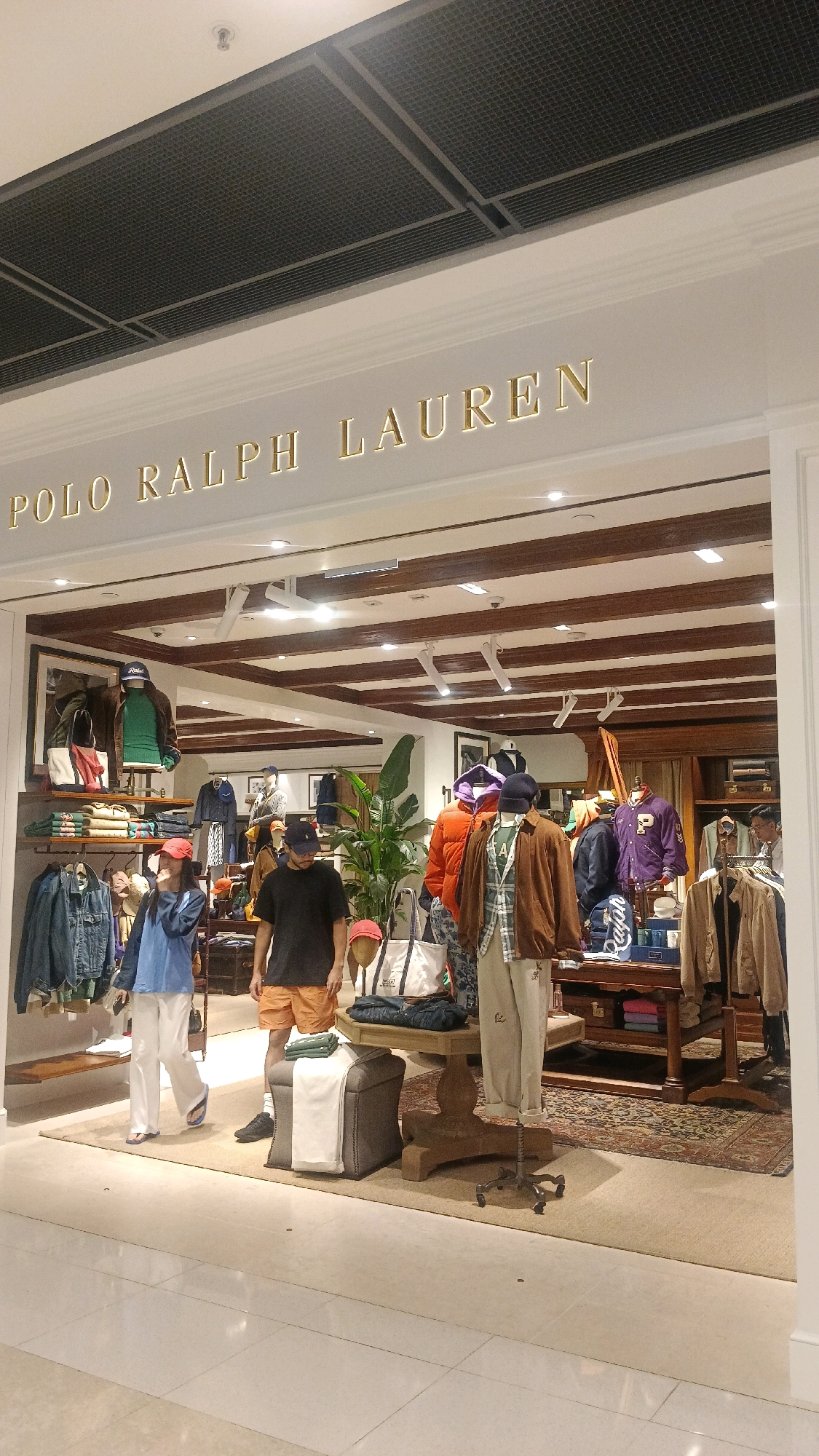 The image shows the entrance to a Polo Ralph Lauren store. The store's name is displayed in gold lettering above the entrance. Inside the store, mannequins are dressed in clothing, and there are racks of clothes on display. The store appears to be well-lit and organized. The flooring appears to be polished concrete or marble. A partial view of a ceiling with metallic grilles and lighting fixtures is visible at the top of the image. The overall impression is that of a clean, upscale retail environment.