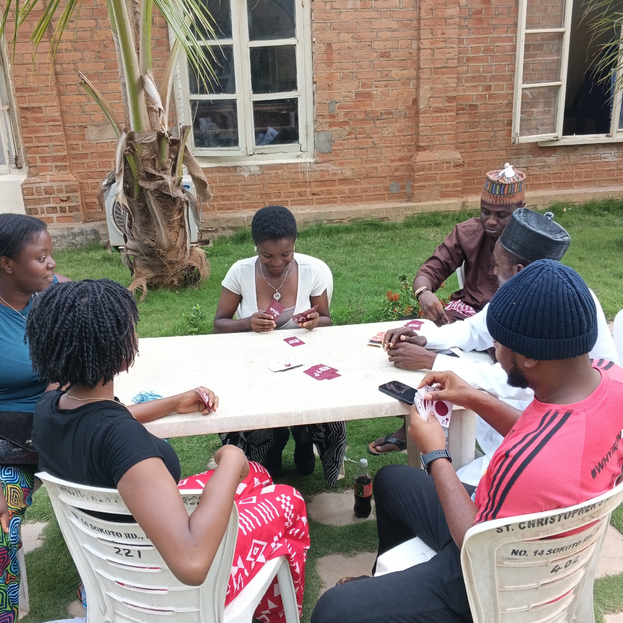 Six individuals are gathered around a light-colored, rectangular table, seated on plastic chairs, engaged in playing a card game in an outdoor courtyard. The setting appears to be the city of Unguwar Sarki, Nigeria, suggested by markings on a chair. A brick building with windows forms the backdrop.
In the foreground, a person in a red shirt with black stripes, wearing a dark beanie, holds a fan of cards, with "12" visible on one card. To their left, a person in a black t-shirt is seated, their arm resting on the table, holding a card. To the left of them, a woman in a teal shirt looks towards the table, while a woman with braided hair in a black t-shirt is also seated, holding a card. Across the table, a woman in a light grey shirt smiles as she looks at her cards, with a necklace featuring a heart pendant. Behind her, a man in a brown traditional outfit and a colorful cap is seated, and next to him, a man in a black cap and white shirt also appears to be participating.
Several decks of cards are spread on the table. A bottle of Coca-Cola and a mobile phone are also present on the table. The ground is covered in green grass, with a paved area around the table. A palm tree with dry fronds is visible to the left of the frame, and an air conditioning unit is mounted on the wall behind it. The overall atmosphere suggests a casual social gathering.