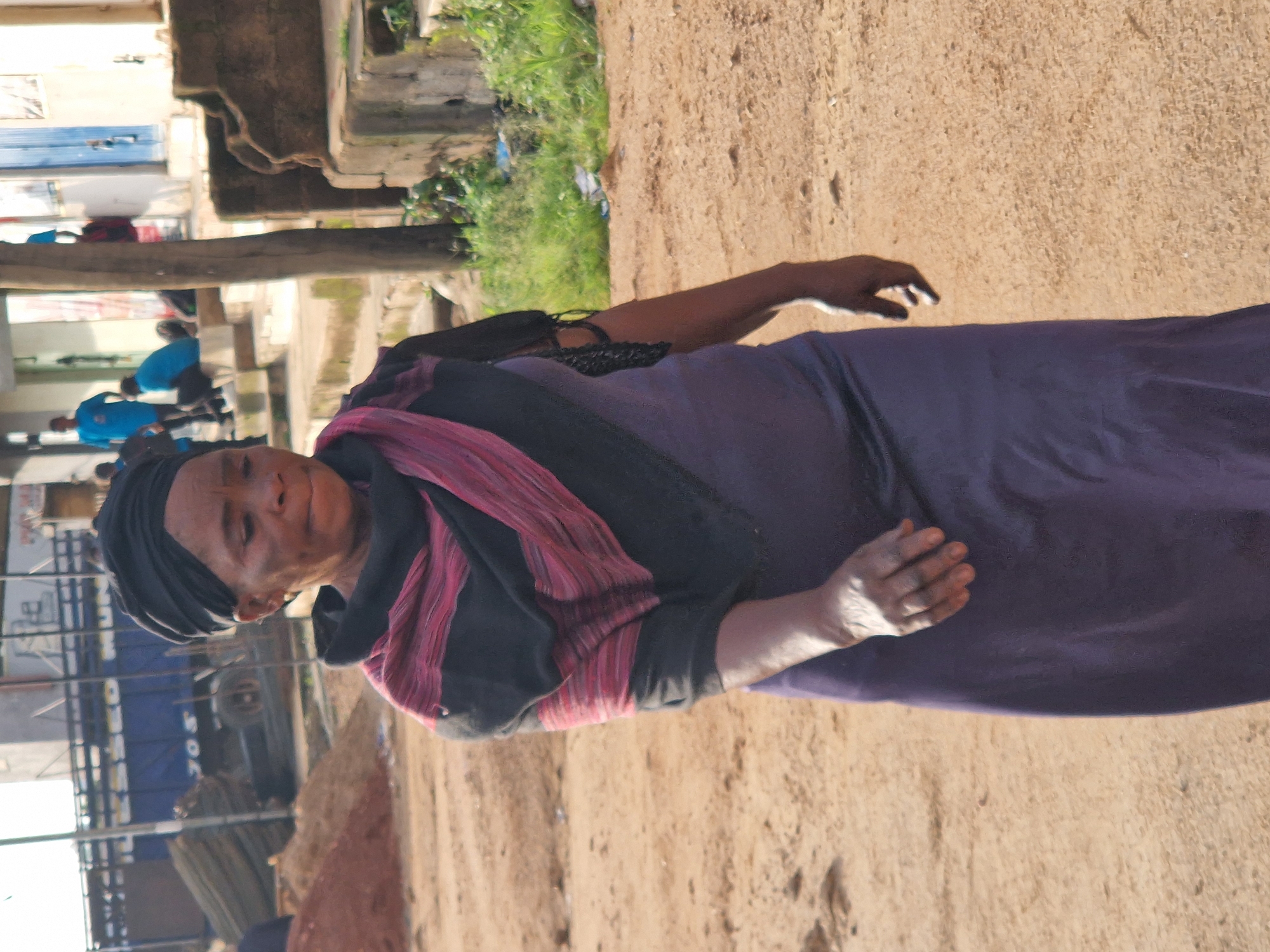 The image captures an outdoor scene in Jos, Nigeria. The primary subject is a woman wearing a dark headscarf, a black top with a pink and red striped scarf draped over it, and a long purple dress. Her head is tilted slightly, and her eyes are closed, suggesting a moment of rest or reflection. Her skin appears to have some texture, and she has a subtle smile. Her right hand is visible, resting in her lap or on the fabric of her dress.
In the background, several other people are present, some wearing blue shirts, and others are less distinct. There are structures that appear to be market stalls or buildings with metal frameworks and corrugated roofing. The ground is a sandy, earthy texture, typical of outdoor environments in such regions. Green vegetation is visible in patches, suggesting some areas are unpaved or have managed to grow. The lighting suggests it is daytime, likely with bright sunlight given the contrast and shadows.
The scene appears to be a bustling environment, possibly a market or a busy street. The overall impression is one of everyday life in a Nigerian urban or semi-urban setting. No specific text is clearly discernible in the image. The activity is static; the focus is on the woman and the general atmosphere of the location.