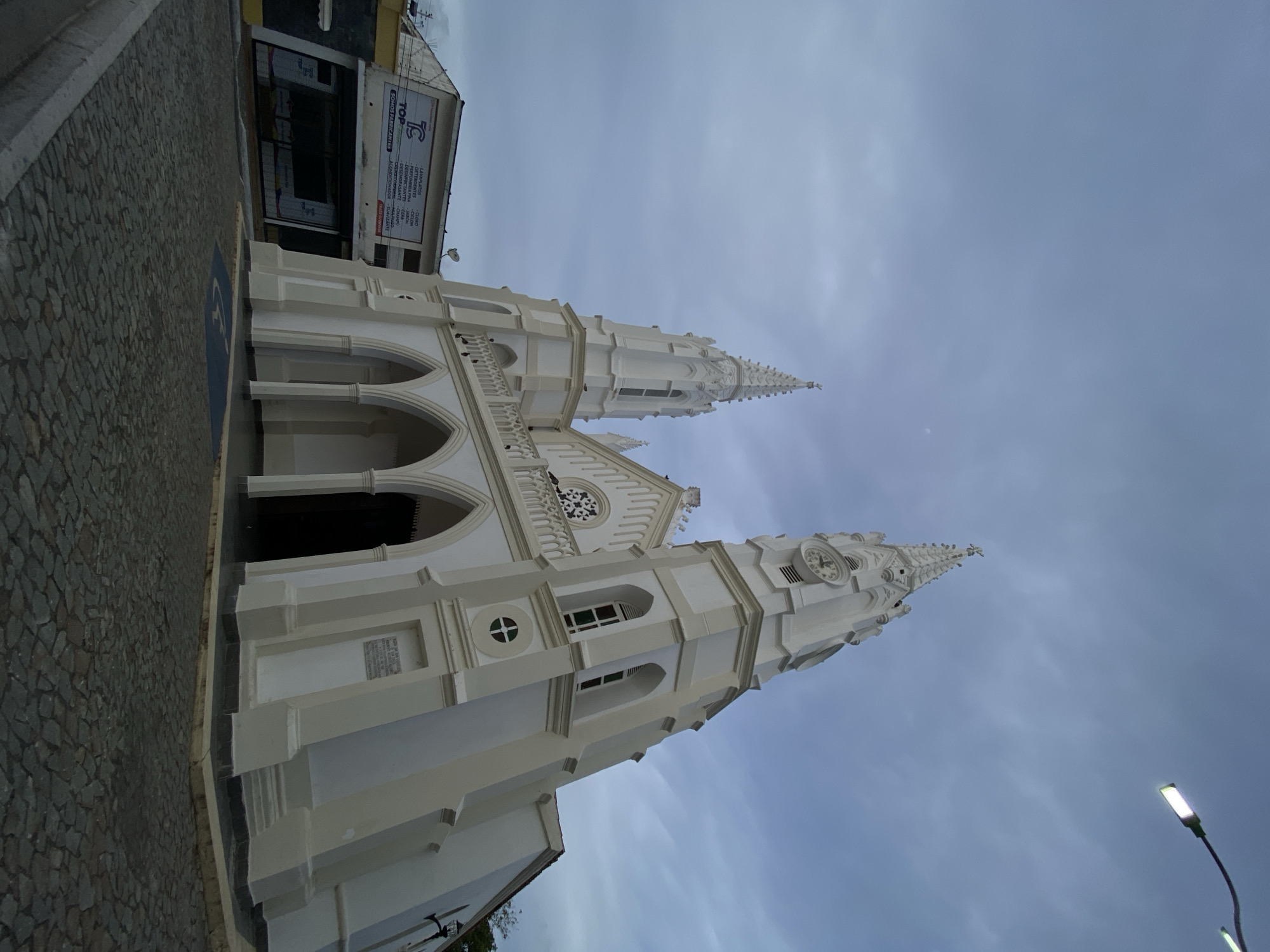 The image displays the Iglesia de San Juan Evangelista (Church of Saint John the Evangelist) in Juan Griego, Venezuela. The central subject is a large, white, Gothic-revival style church with two tall spires, both topped with crosses. The right spire features a visible clock face. The church's facade includes multiple pointed arches forming a portico, a balustraded balcony, and a circular rose window above the main entrance. Various arched and round windows are present along the building's white walls, which feature beige trim.
The ground immediately in front of the church is paved with dark grey cobblestones. A blue square with a white international accessibility symbol is painted on the cobblestones near the church's entrance. To the left of the church, a two-story commercial building is visible. This building has a white upper facade with a prominent "TOP" logo and a list of products including "LAVAPLATOS," "DETERGENTES," and "PERFUMERIA." Black-framed windows are visible on the lower level, and a satellite dish is mounted on the building's exterior. A street light with a luminous LED fixture extends into the upper right portion of the frame. The sky is uniformly overcast, and no individuals or vehicles are present in the scene.