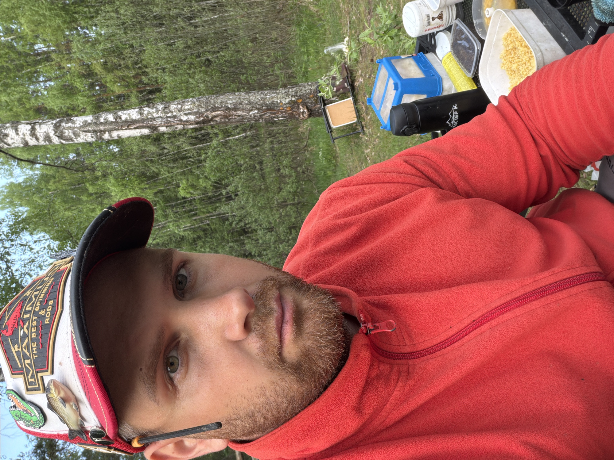 The image shows a man with light skin and a beard, wearing a red fleece jacket and a baseball cap adorned with fishing-related emblems (a fish, an alligator, and text "MAXIMA THE BEST SPINNING & RODS"). He appears to be outdoors. In the background, there's a birch tree and a forest scene suggesting a rural or wilderness area. To his right, there is a table with fishing gear and accessories including containers and small items. It is not possible to determine whether the location is in the city of Chaykovsky, Russia based on the image.