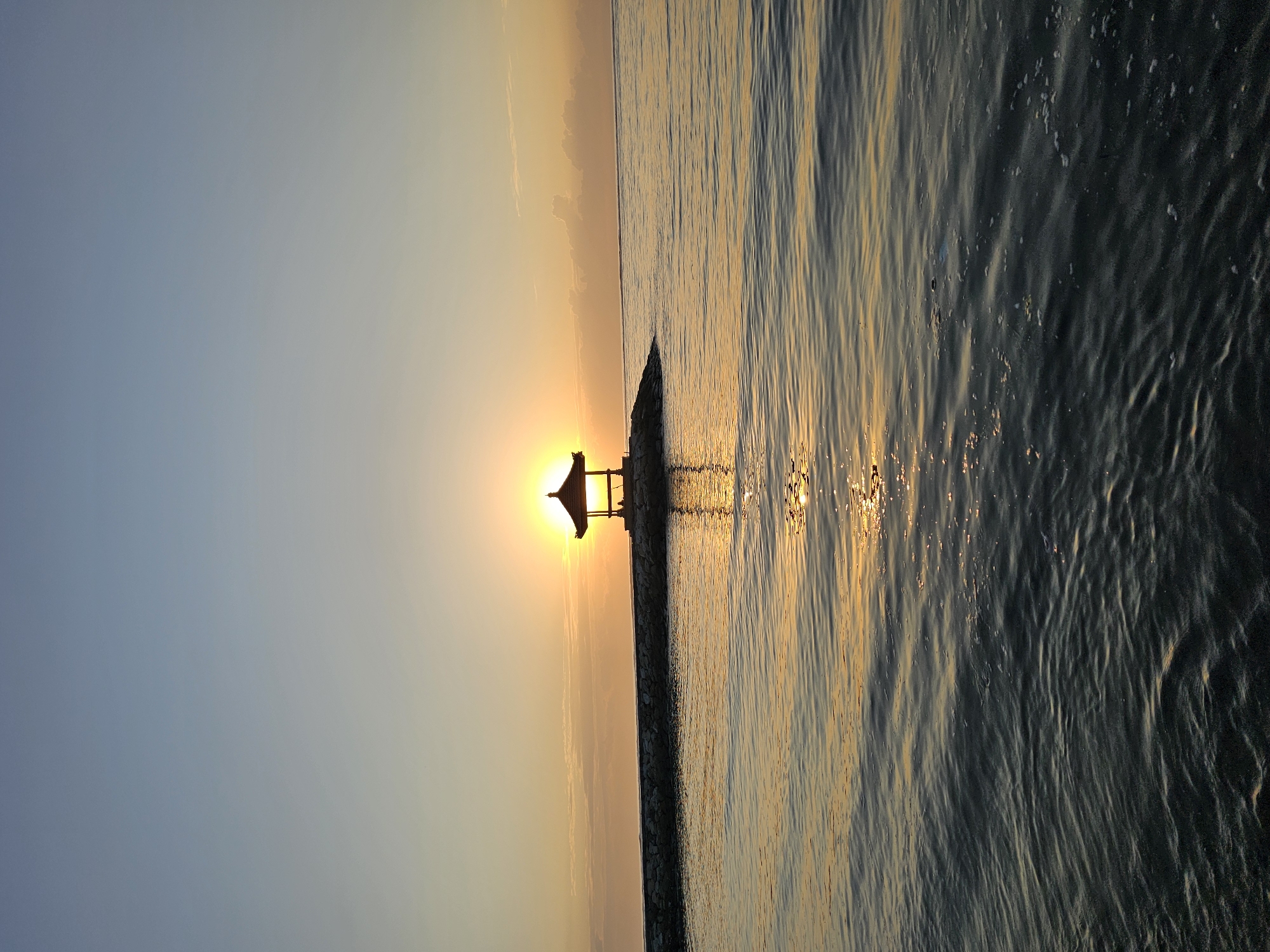 A serene scene unfolds at Sanur Beach in Denpasar, Indonesia, as the sun dips below the horizon, casting a warm, golden glow across the water. The sky transitions from a soft, pale blue at the top to a vibrant orange hue near the horizon, painted with wispy clouds. In the middle ground, a small, dark gazebo sits silhouetted on a rocky jetty that juts out into the calm sea. The sun is positioned directly behind the gazebo, its rays emanating outwards, creating a halo effect. The water's surface is gently rippled, reflecting the warm colors of the sunset and the structure on the jetty. The overall impression is one of tranquility and natural beauty.