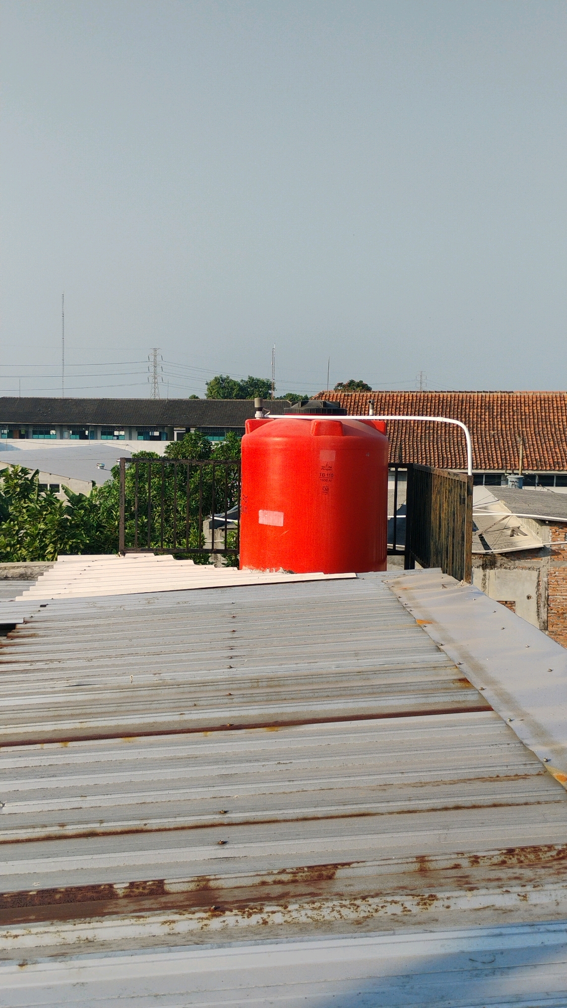 The image shows a rooftop scene in Gumpang, Indonesia, dominated by a large, bright red water tank. The tank is cylindrical with a rounded top and is positioned on a lower rooftop that appears to be made of corrugated metal. The corrugated metal rooftop in the foreground is weathered and shows signs of rust. Behind the water tank, there is a dark metal fence and lush green trees. Further in the background, there are buildings with tiled roofs, some with a dark, flat roof structure, and in the distance, utility poles and transmission towers are visible against a hazy, pale blue sky. The overall impression is of an industrial or semi-urban environment.