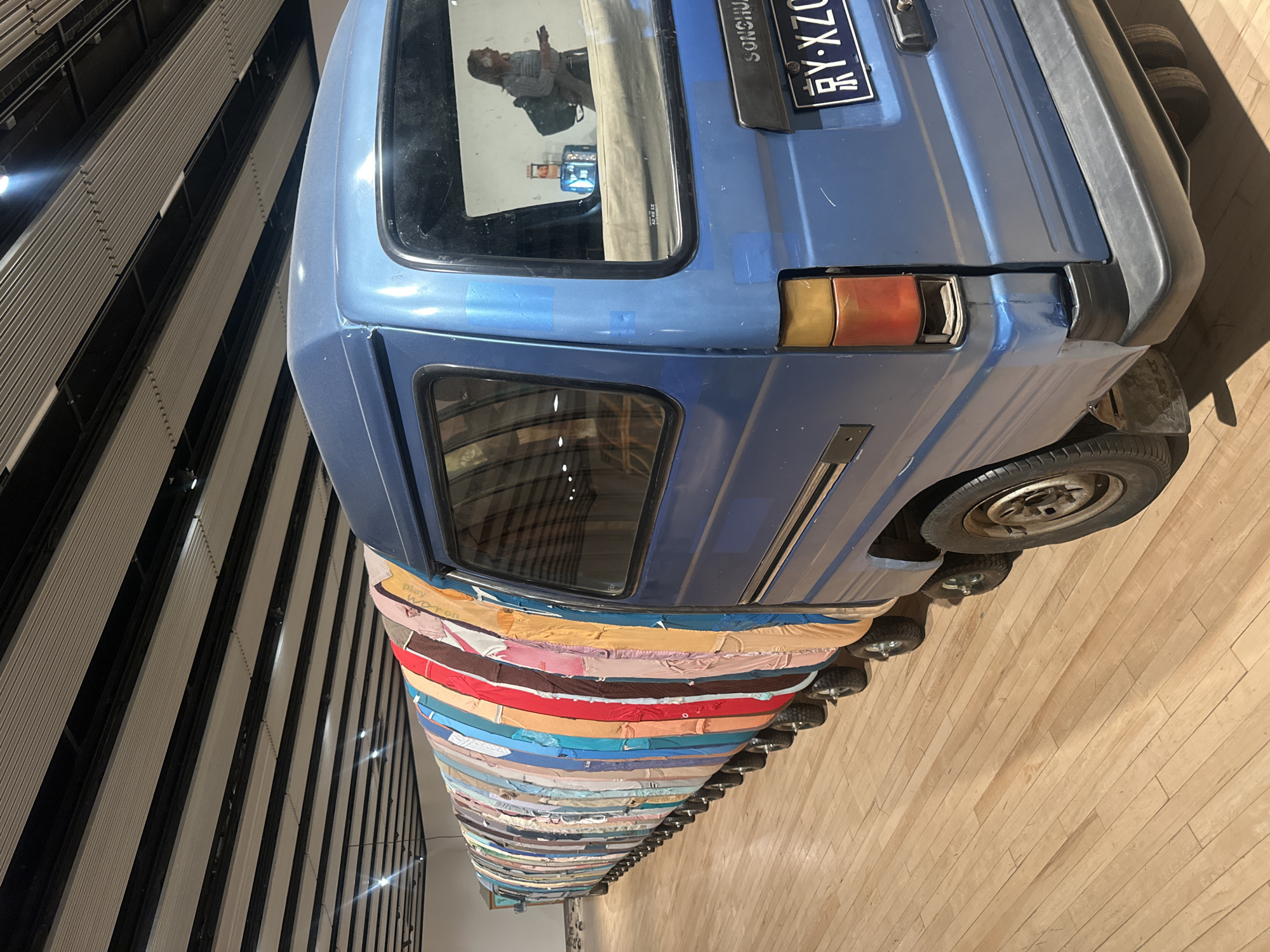 The image shows an art installation, "When Attitudes Become Form: Bernar Venet’s Pile of Coal," inside the Hayward Gallery, part of the Southbank Centre in London. The installation consists of a blue van from which a long, cone-shaped structure extends, made up of stacks of multi-colored mattresses. The van has a license plate visible, reading "RY-XZ07X-TAIWAN." The mattresses are tightly packed and create a visually striking pattern. The van is positioned on a wooden floor. In the van's rear window, a reflection is visible, showing a person holding a mobile phone. A section of the gallery's wall, with a series of black and white vertical panels, is visible in the background.