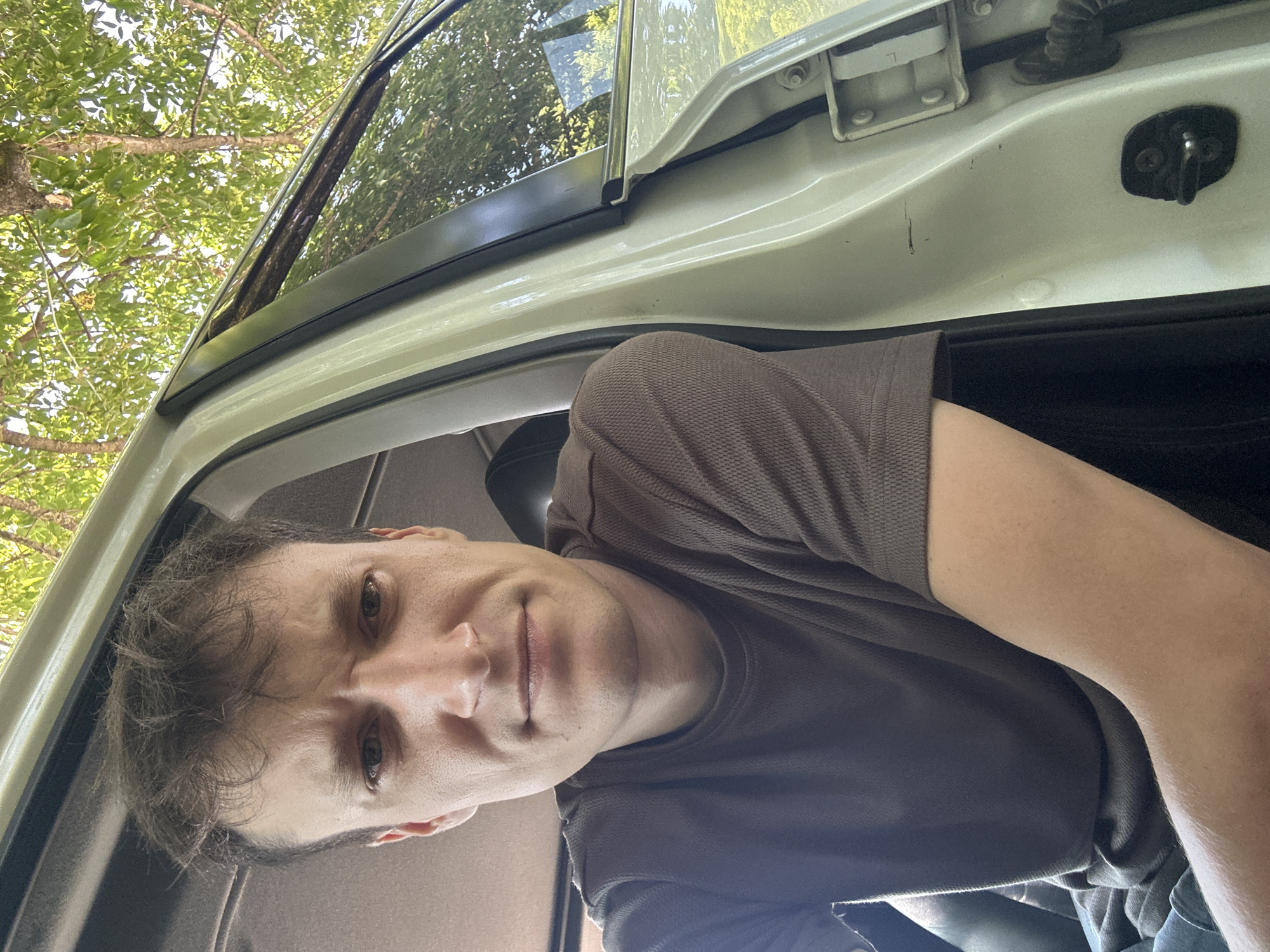 The image is a close-up, selfie-style shot of a young adult male sitting inside a vehicle. He is wearing a dark brown t-shirt and appears to be looking directly at the camera with a neutral expression. His hair is brown and somewhat messy. The interior of the car, including the door frame and a portion of the door panel, is visible. Outside the car, through the open door and window, trees and foliage can be seen, suggesting an outdoor, possibly rural or suburban setting. The lighting suggests it is daytime. There is no specific indication of the location being in Makiv, Ukraine.