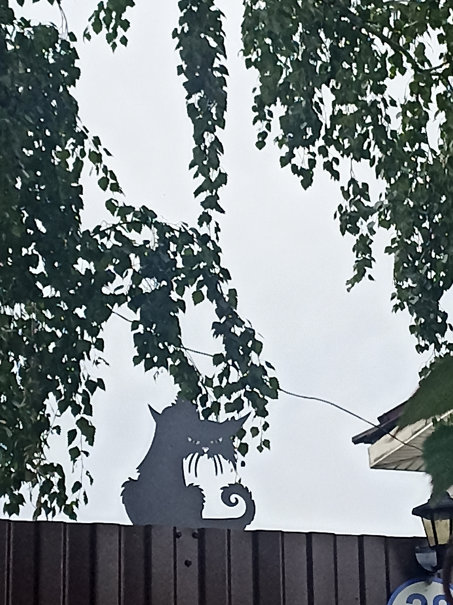 The image shows a black silhouette of a menacing-looking cat perched on top of a dark brown wooden fence. The cat's silhouette is detailed, with sharp ears, an open mouth showing teeth, and a coiled tail. Behind the fence, the silhouette of the cat is framed by the branches of a leafy tree, with dark green leaves hanging down against a pale, overcast sky. To the right of the cat, a corner of a light-colored roof with dark trim is visible, and a black lamppost with a yellowish light fixture stands on the fence. In the lower right corner, a circular blue sign with white numbers "20" is partially visible. The overall scene appears to be an outdoor decorative element in a residential or garden setting.