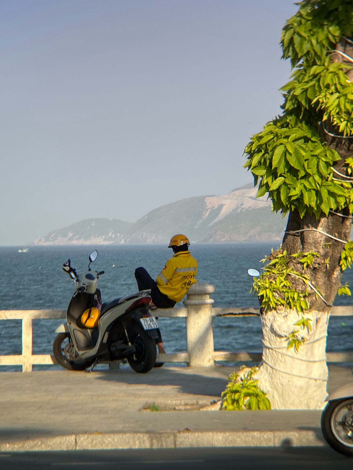 The image depicts a person wearing a yellow jacket and helmet sitting on a railing overlooking the ocean in Nha Trang, Vietnam. A scooter is parked next to them. The background shows a hazy coastline with mountains and a calm sea. The foreground is a concrete sidewalk and road. The overall atmosphere is serene and peaceful, with the person appearing to be taking a break and enjoying the view. The presence of the scooter suggests they might be a delivery person or a tourist. The yellow helmet and jacket, and the scooter's license plate containing Vietnamese characters, further confirm the location.