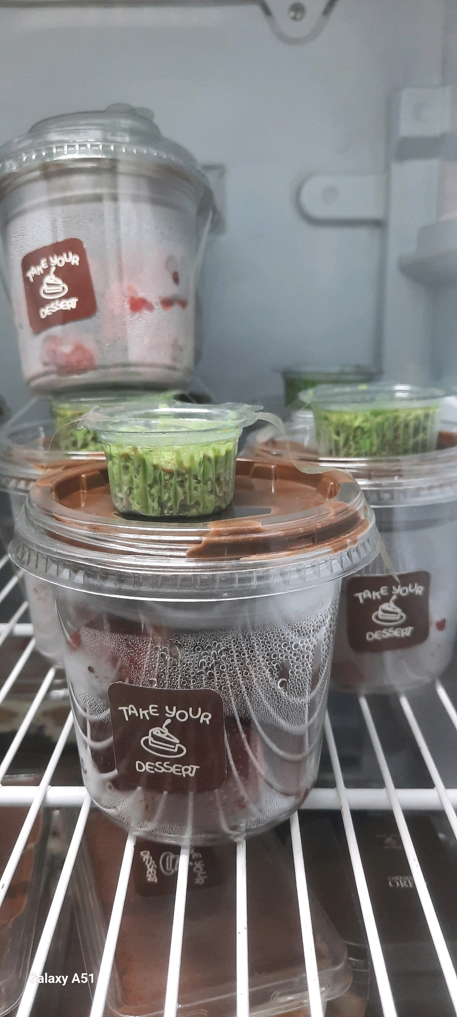 The image shows several clear plastic containers of desserts inside a refrigerator. The containers are stacked on white wire shelves. The desserts appear to be layered, with some containing a brown base and others a green topping. Some containers have labels that read "TAKE YOUR DESSERT" with a cake graphic. Condensation is visible on the outside of some containers. The environment appears to be a domestic refrigerator. The location in the city of Beru, Indonesia cannot be determined from the image.
