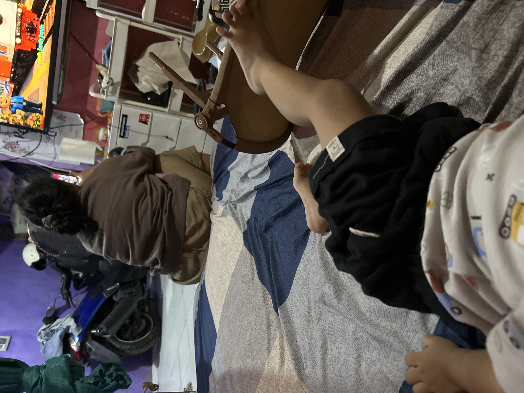 In a room in Depok, Indonesia, a low-angle, close-up shot shows a child's legs and feet lying on a bed with a patterned comforter. The child is wearing black shorts and a light-colored onesie with colorful cartoon illustrations of vehicles. To the right of the child's legs is a brown plastic child's rocking chair. In the background, a person is seated facing away from the camera, wearing a brown sweatshirt and tan pants, looking at a television screen displaying a cartoon. A blue motorcycle with a white helmet on its handlebars is parked to the left. The room's walls are painted purple and pink.