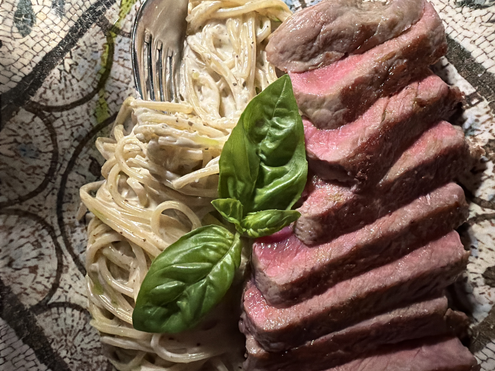 A close-up, top-down shot showcases a plate of food. On the left side, there is a serving of spaghetti coated in a creamy sauce, with a fork resting on top of the pasta. In the center, a sprig of fresh basil is placed. To the right, there is a stack of sliced medium-rare steak. The plate itself features a mosaic-like design in neutral tones. The location is indicated as Rafailovići, Montenegro, but there is no other specific information regarding the environment.