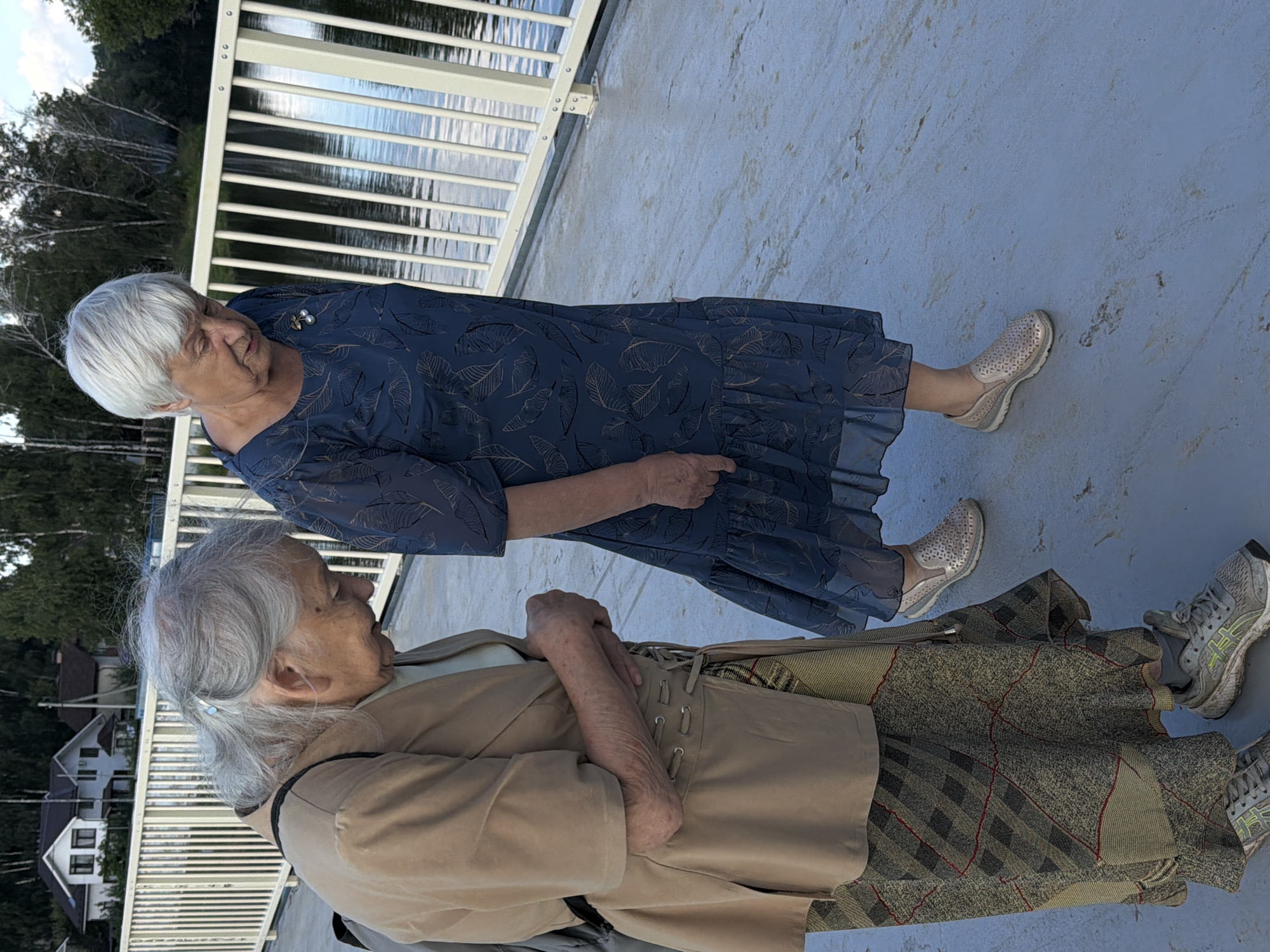 Two elderly women are standing on a bridge with a white railing. The woman on the left is wearing a tan jacket over a white shirt and a patterned skirt, and has gray hair. The woman on the right is wearing a blue patterned dress and has short white hair. They are both looking down. The background shows a body of water and trees, with houses visible in the distance. The bridge appears to be in a park or recreational area in the city of Udelnaya, Russia.