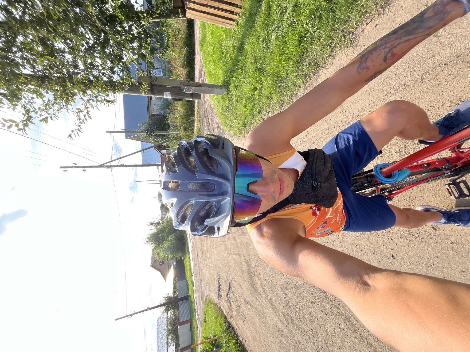 The media file shows a person riding a bicycle outdoors. The subject is wearing a blue cycling helmet with reflective rainbow-tinted glasses, an orange and white cycling jersey, and blue shorts. They have a tattoo on their left arm. The scene is a rural or suburban area with a dirt road, green grass, trees, and some buildings in the background. There are utility poles and wires visible, suggesting a populated area. The sun is shining brightly, indicating it is daytime, likely late morning or afternoon, and the weather appears to be good. The perspective is from the rider's point of view, as if it's a selfie taken while cycling. The overall mood seems to be active and recreational. No discernible text is visible in the image. The location context is Chaikovsky, Russia.