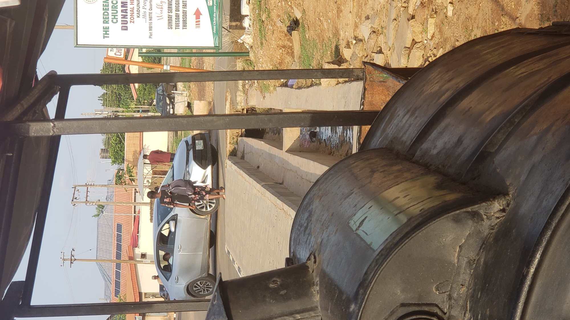 The image, taken in Narayi, Nigeria, captures a daytime street scene from a slightly elevated perspective, possibly from inside a vehicle or structure. In the immediate foreground on the right, a large, dark, possibly plastic, water tank is partially visible.
The midground features a paved road with concrete curbs and drainage structures running alongside it. Several people are present: a woman wearing a floral dress and a dark top, carrying a backpack, stands next to a silver sedan on the left. Behind her, a white motorcycle is partly visible. Further down the road, another person in a red top stands near a second silver car.
In the background, various buildings with reddish-brown roofs are visible, one of which appears to have solar panels installed. Utility poles with overhead power lines line the street. A prominent, partially obscured sign for "THE REDEEMED CHURCH" is in the upper background. The sign also mentions "DUNAMI," "ZONAL HEADQUARTERS," and "KADUNA," along with "Plot MB16 NBTE Quarters." It lists "Weekly" services: "SUNDAY SERVICE," "TUESDAY DIGGING," and "THURSDAY FAITH," concluding with the partial phrase "Jesus Christ the same yesterday..." The license plate "FGE 021 B" is partially visible on one of the silver cars.
The weather is bright and sunny with clear skies.