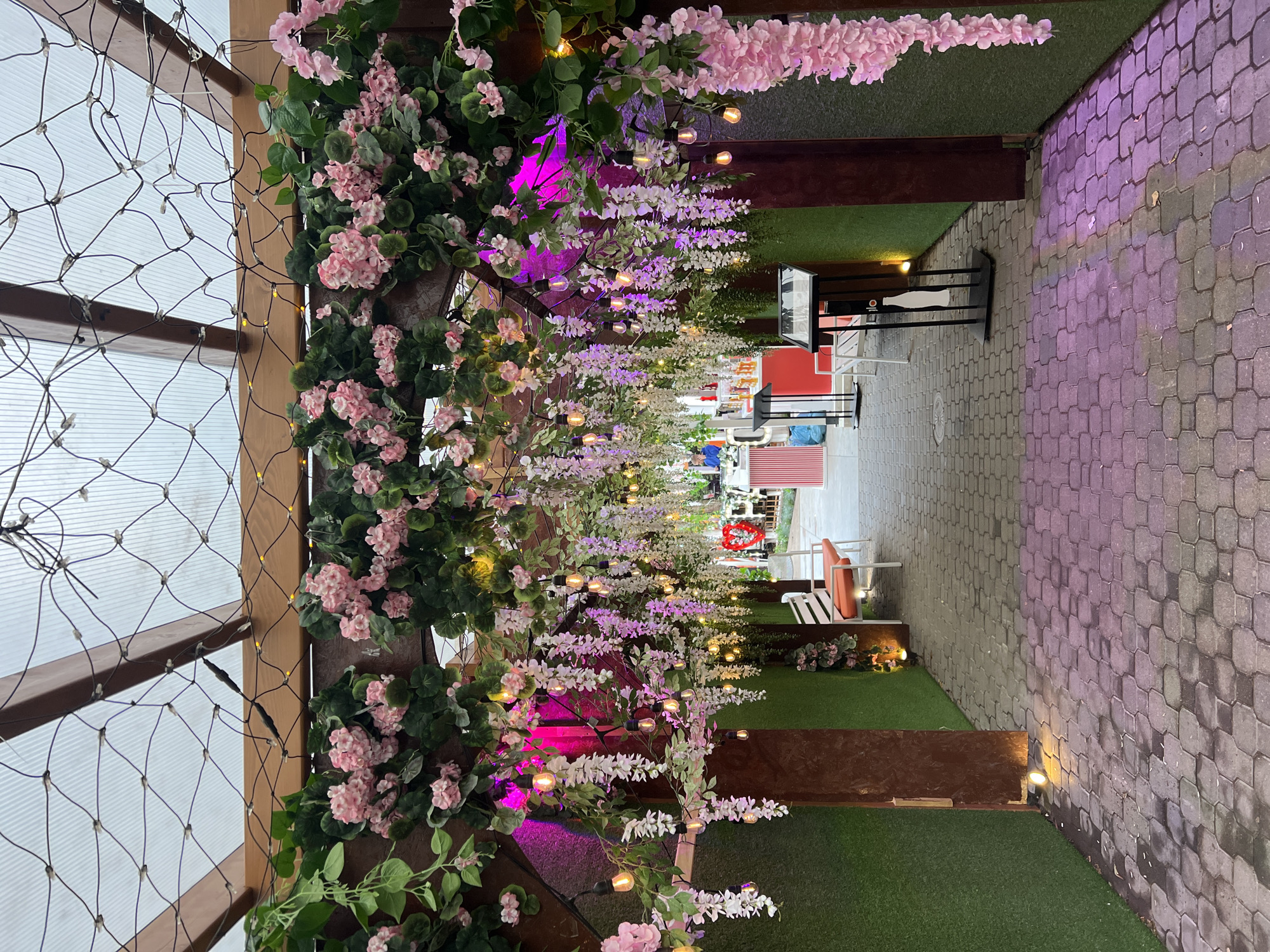 This image depicts an outdoor seating area, likely a restaurant or cafe, decorated with artificial pink and white wisteria flowers and string lights under a clear, corrugated roof. The pathway is paved with gray interlocking bricks, some of which are illuminated by pink and yellow lights. Green artificial turf lines the walls on either side. In the background, there are white benches with orange cushions, tables, and a red and white striped structure that might be a counter or bar. The scene appears to be set in Białystok, Poland, based on external information. There are no people clearly visible in the foreground.