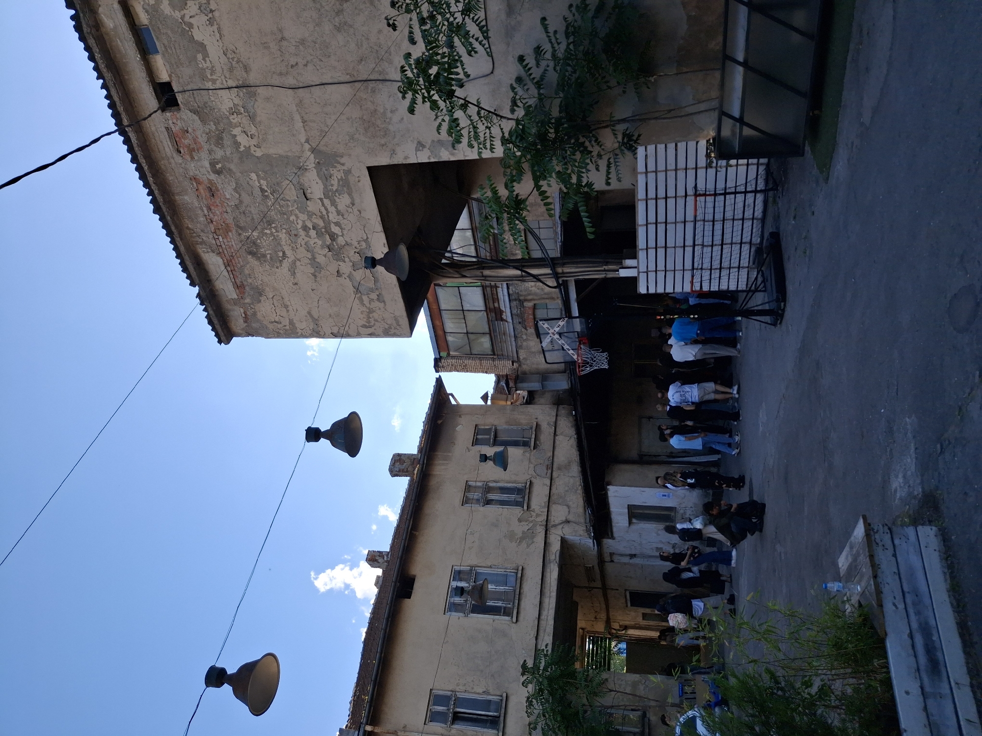 The image shows an outdoor courtyard in an urban environment, likely in Unknown, Unknown. The space is bordered by buildings with aged facades, featuring multiple windows. A row of people is standing, possibly lining up, near what appears to be a basketball hoop and backboard. A second group is seated further back in the courtyard. Three vintage-style lamps are suspended overhead, and power lines crisscross the blue sky visible between the buildings. Greenery is present, adding a touch of nature to the otherwise urban scene.