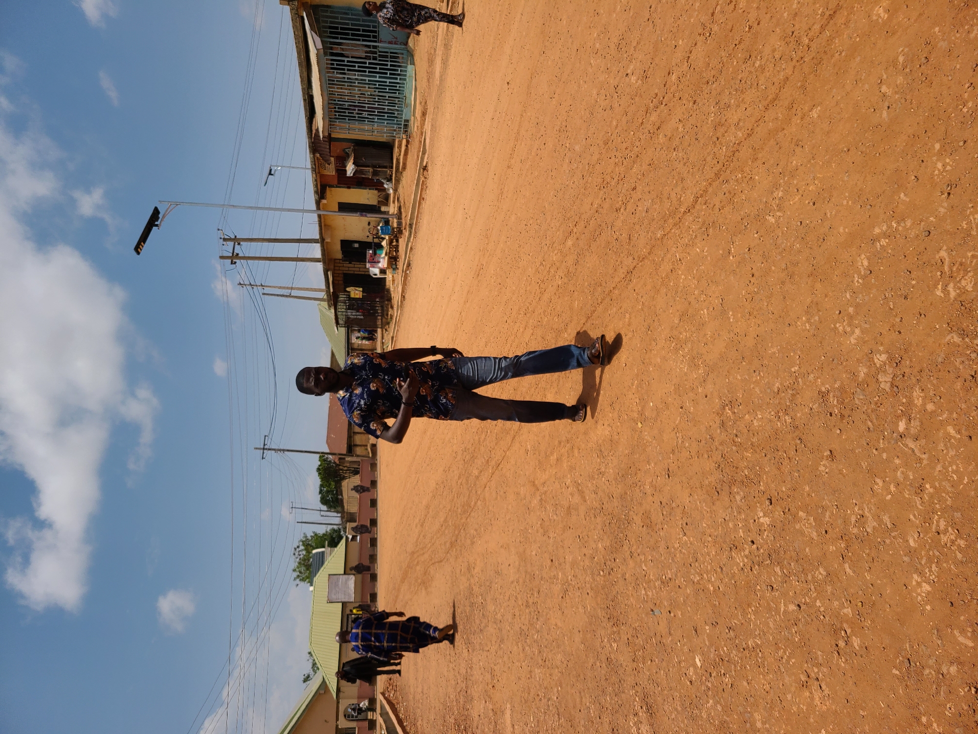 The image shows a street in the city of Unknown, Unknown. The street is unpaved and covered with reddish-brown dirt. A man wearing a patterned shirt and jeans is standing in the middle of the street. There are buildings on either side of the street, with utility poles and power lines running along the side. Other people can be seen in the distance, walking on the street. The sky is blue with some clouds.