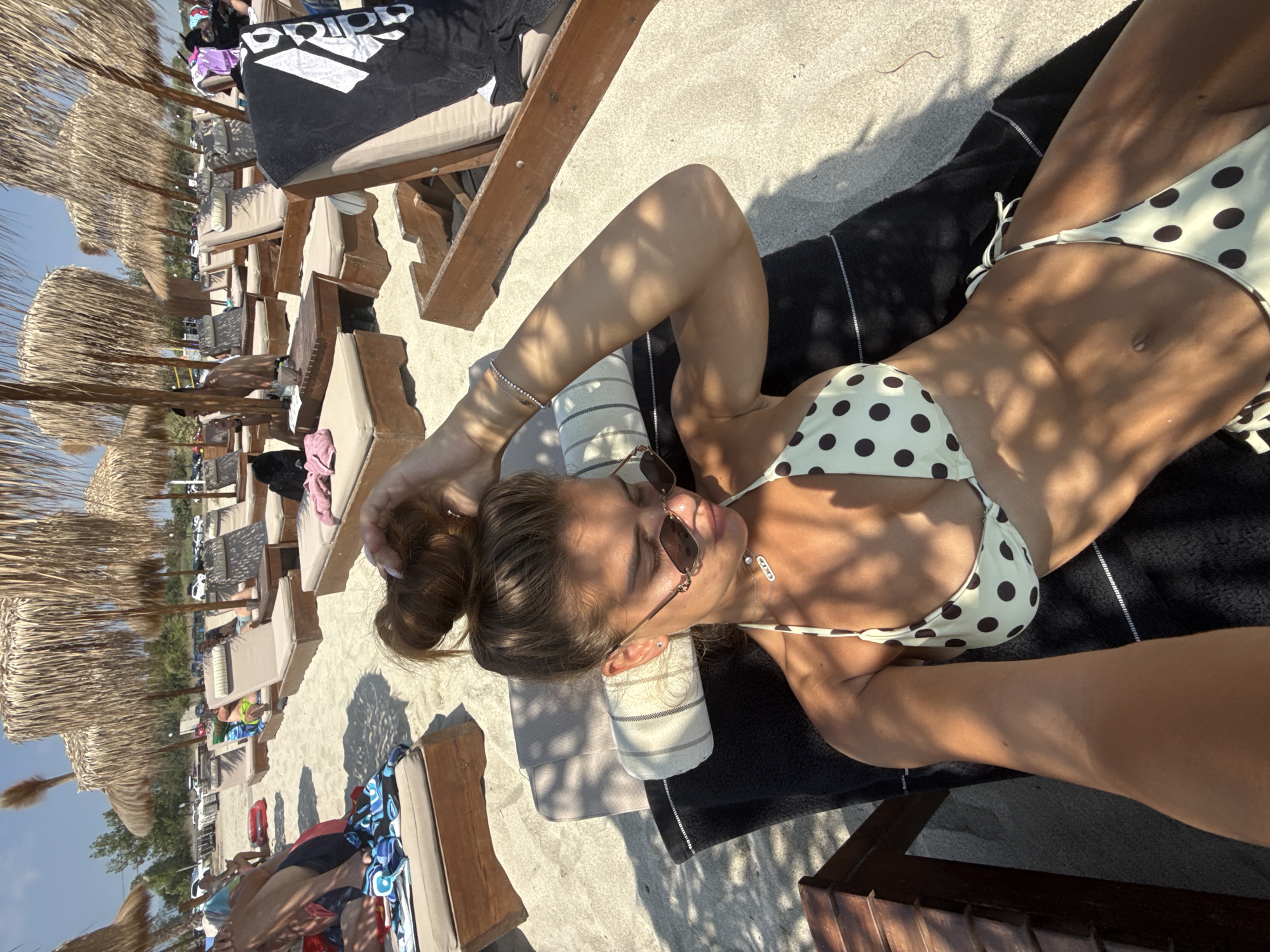 A young woman with her hair in a bun is lying on a beach chair on a sandy beach. She is wearing sunglasses and a bikini with a polka dot pattern. She is resting her arm behind her head and appears to be relaxing. In the background, there are more beach chairs, umbrellas, and other people on the beach. The scene suggests a sunny day at a beach resort. The location is the Olympic Beach, part of Katerini Municipality, Greece, known for its organized beaches and tourist facilities.