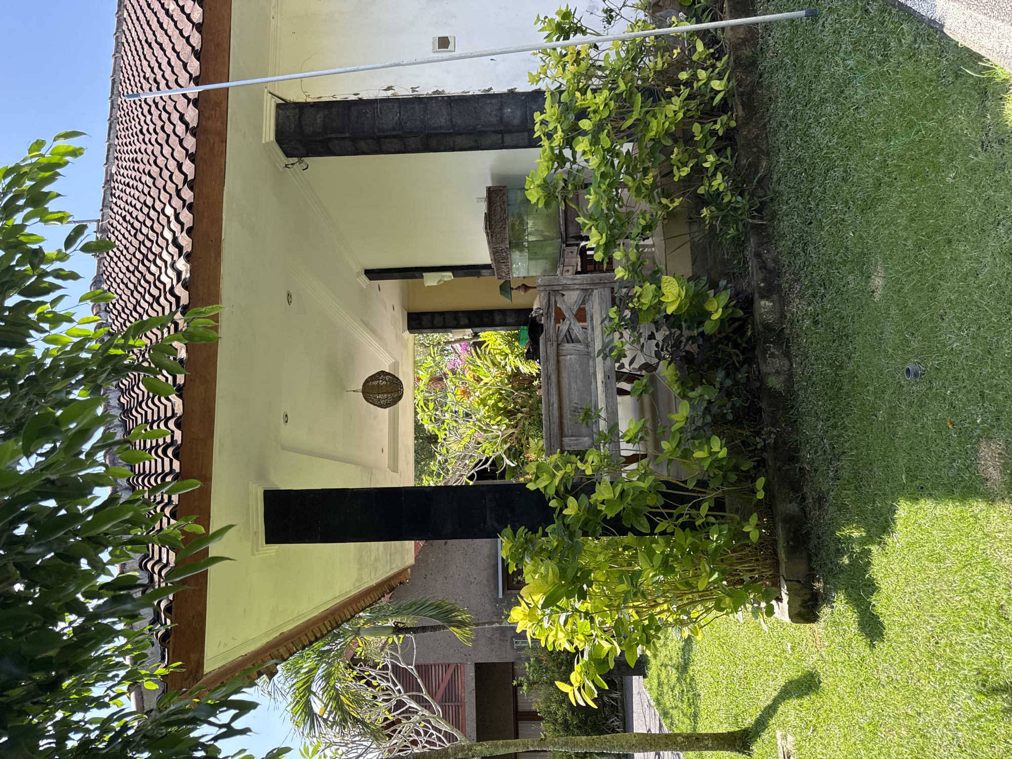 The image depicts a residential scene in Kuta Selatan, Indonesia. The architecture features a building with a tiled roof and white exterior walls. Dark-colored support beams are visible. Lush greenery and plants are growing around the building and in the adjacent yard. The yard is covered in green grass. A paved walkway is present on one side of the yard. A nearby building with brown shutters is visible in the background. The sky is blue and clear. There are no people visible in the image.