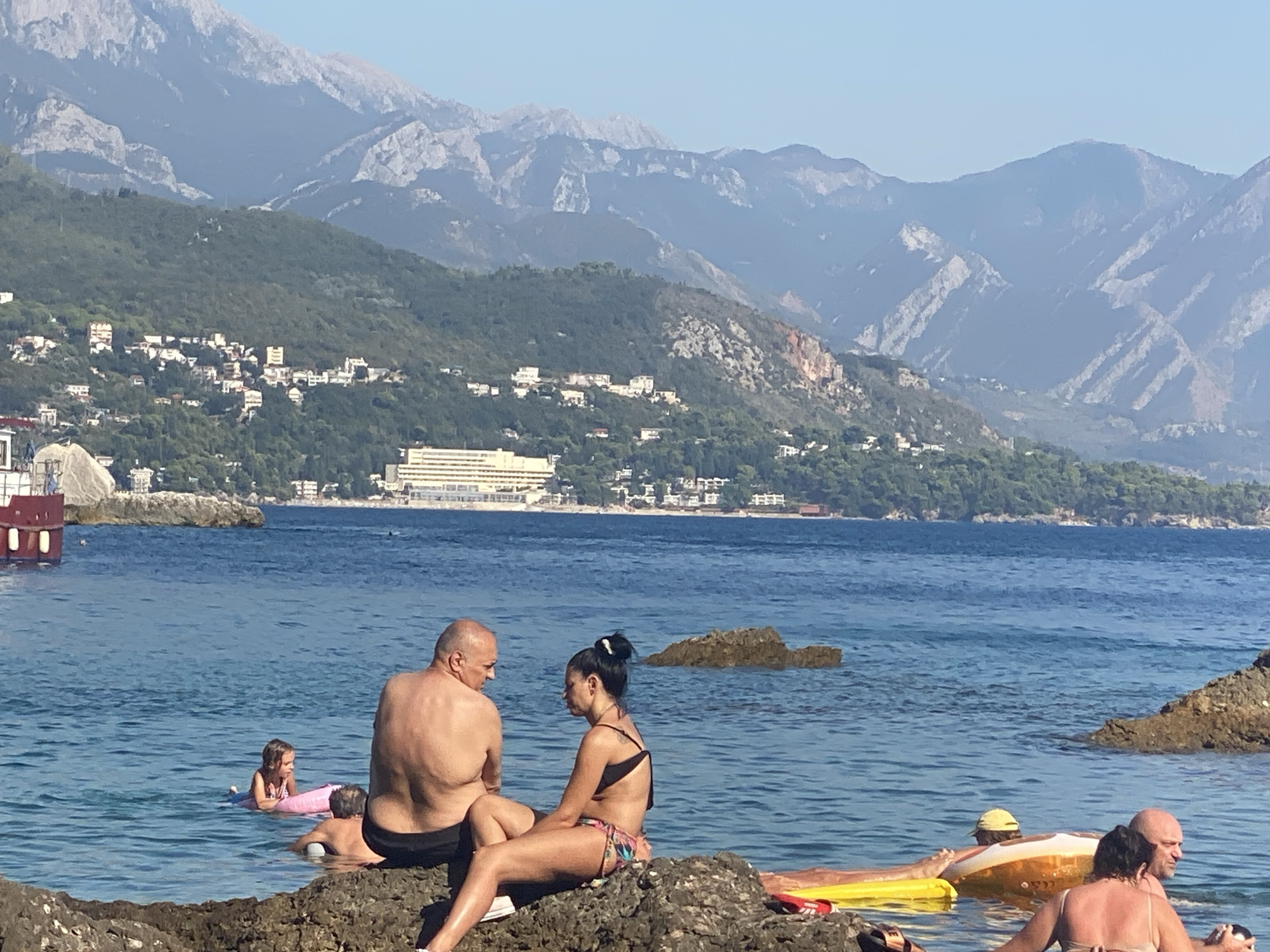 The image captures a serene beach scene in Sutomore, Montenegro, bathed in the warm glow of daylight. The sky is a clear, unblemished blue, suggesting fair weather. In the foreground, a couple is seated on a rocky outcrop by the water's edge. The man, bald and with a robust build, faces away from the camera, engaged in conversation with the woman beside him. She, with her dark hair tied up, is dressed in a colorful bikini.
Further out in the calm, blue sea, several other individuals are enjoying their day. A young girl is on a pink inflatable ring, looking towards the shore. Another person is partially visible in the water, wearing a yellow cap. A bright yellow inflatable float and an orange and white one are also present in the water, indicating leisurely water activities.
In the mid-ground, the coastline is dotted with buildings, ranging from small houses to larger hotels, nestled amongst lush green hillsides. A prominent, long, white building, likely a hotel, stands out on the elevated terrain. In the background, majestic, rugged mountains with visible rock formations rise up, their peaks softened by distance and the haze of the day. The overall atmosphere is one of relaxation and summer enjoyment, with the natural beauty of the coastline and mountains providing a picturesque backdrop. No visible text is present in the image.