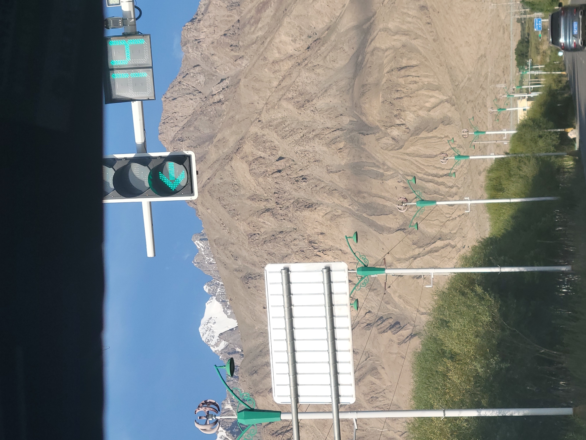 The image shows a view of a road in Tashkorgan, China, with a large mountain range in the background. A traffic light is visible in the foreground, displaying a green pedestrian symbol and a countdown timer showing "14" seconds. There are also several lampposts with decorative tops and a large white rectangular sign mounted on poles. Trees line the side of the road, and a car is driving away from the camera. The sky is clear and blue, and the sun is shining. The mountains are rocky and rugged, with some snow on the peaks. The overall scene suggests a rural or semi-rural area with infrastructure for traffic control.