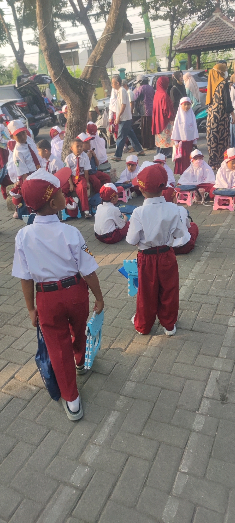 The image depicts an outdoor scene in Banjarsari, Indonesia, featuring a group of young students in uniform. The students are wearing white shirts, red pants, and red and white caps. Some are sitting on the ground, while others are standing. A few are holding blue objects that appear to be folded chairs or learning aids. In the background, there are adult figures, cars, and a tree. The ground is paved with interlocking brick tiles. The overall atmosphere suggests a school-related activity or gathering.