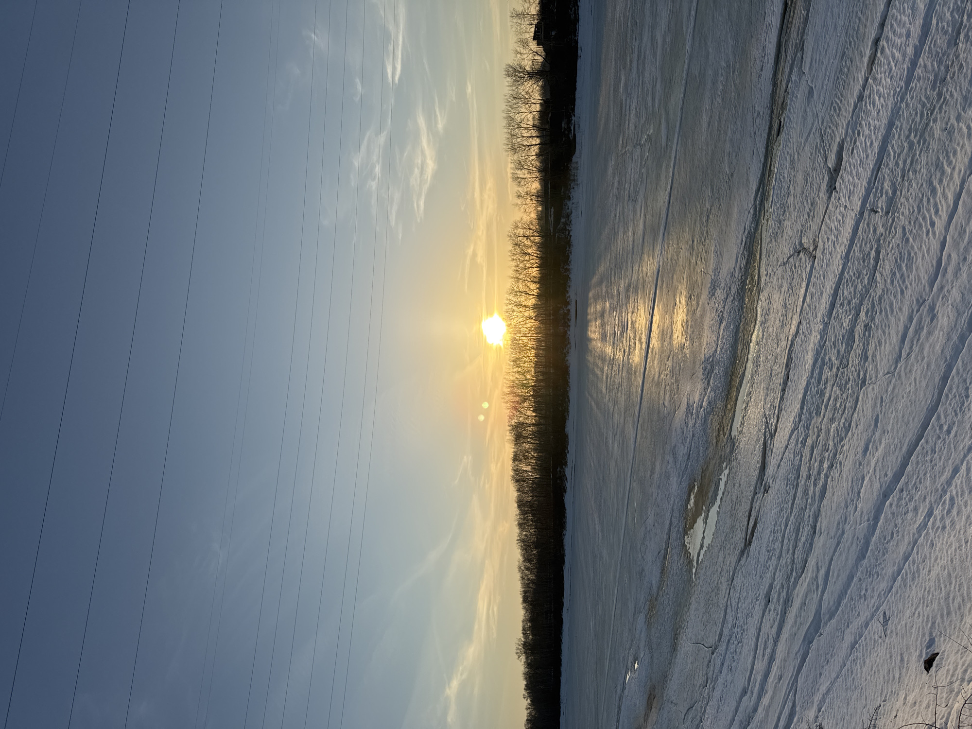 The image depicts a winter landscape near Chaikovsky, Russia. The sky is a gradient of blue to orange-yellow near the horizon, where the sun is setting. Power lines are visible on the left side of the frame. A line of trees separates the sky from a snow-covered field. The snow cover shows patterns of ridges and tracks. The sun's reflection is visible on the snow's surface. There are no people visible in the scene.