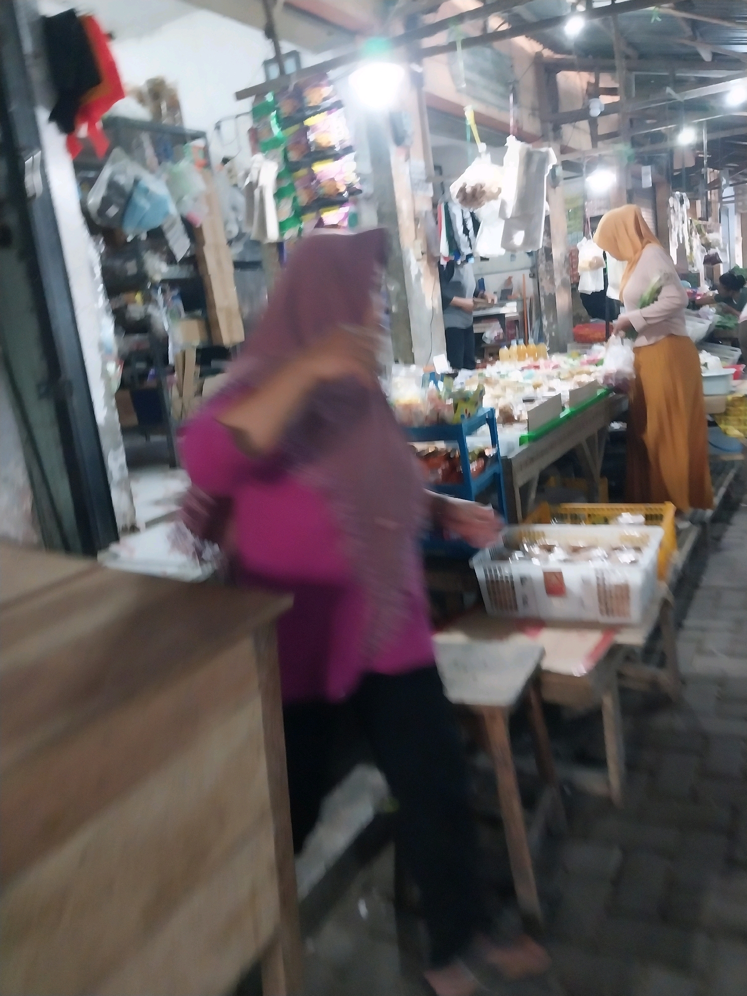 The image depicts a marketplace scene in Penataran, Indonesia. In the foreground, a person wearing a magenta top and black pants is partially visible, with a wooden counter to their left. In the midground, a woman in a light pink top and an orange skirt, with a matching headscarf, is standing and holding a plastic bag. She appears to be interacting with a stall that displays various food items and goods. Further in the background, other individuals are present, and shelves are stocked with merchandise. The environment suggests an outdoor or semi-outdoor market with overhead structures and lighting.