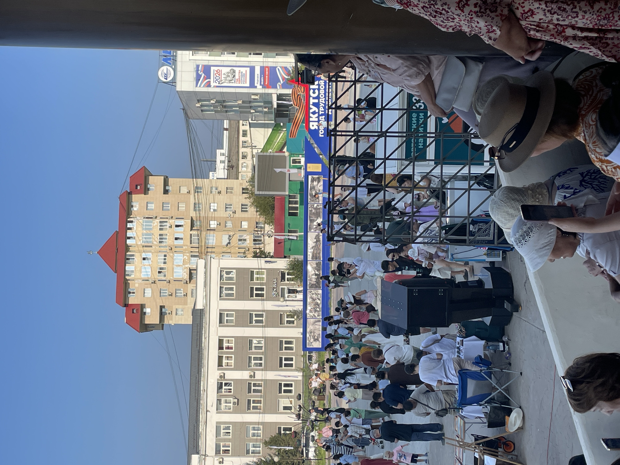 The image shows a crowd of people gathered in a public square in Yakutsk, Russia. A large, light-colored building with a red roof dominates the background, with other multi-story buildings and a banner reading "YAKUTSK" and "GOROD TRUDOVOY SLAVY" (City of Labor Glory) visible. In the foreground, a metal fence separates the viewers from a stage or display area, where tables with goods and folding chairs are set up. People are standing and observing, some appear to be vendors, while others are spectators. The weather is clear and sunny, suggesting a daytime event. The overall scene depicts a community gathering or celebration.