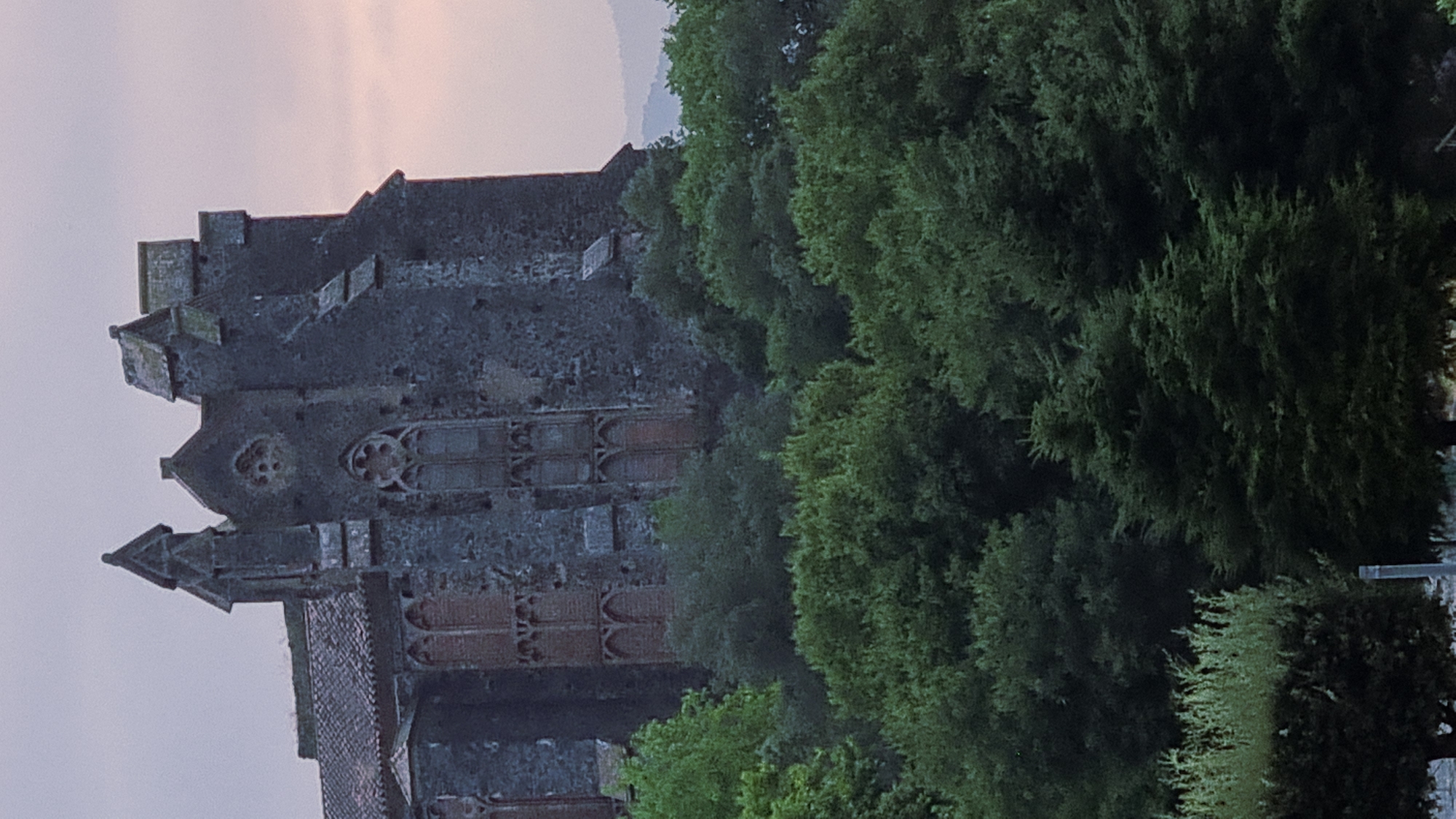 The image shows a view of the Torre Gironella, a medieval defense tower, in Sant Joan Les Fonts, Spain. The tower is constructed of dark stone and features gothic architectural elements, including pointed arch windows and decorative stonework. It is partially obscured by lush green trees in the foreground. The sky above is overcast with a hint of pink, suggesting either dawn or dusk. The overall scene depicts a historical structure set within a natural, verdant environment.