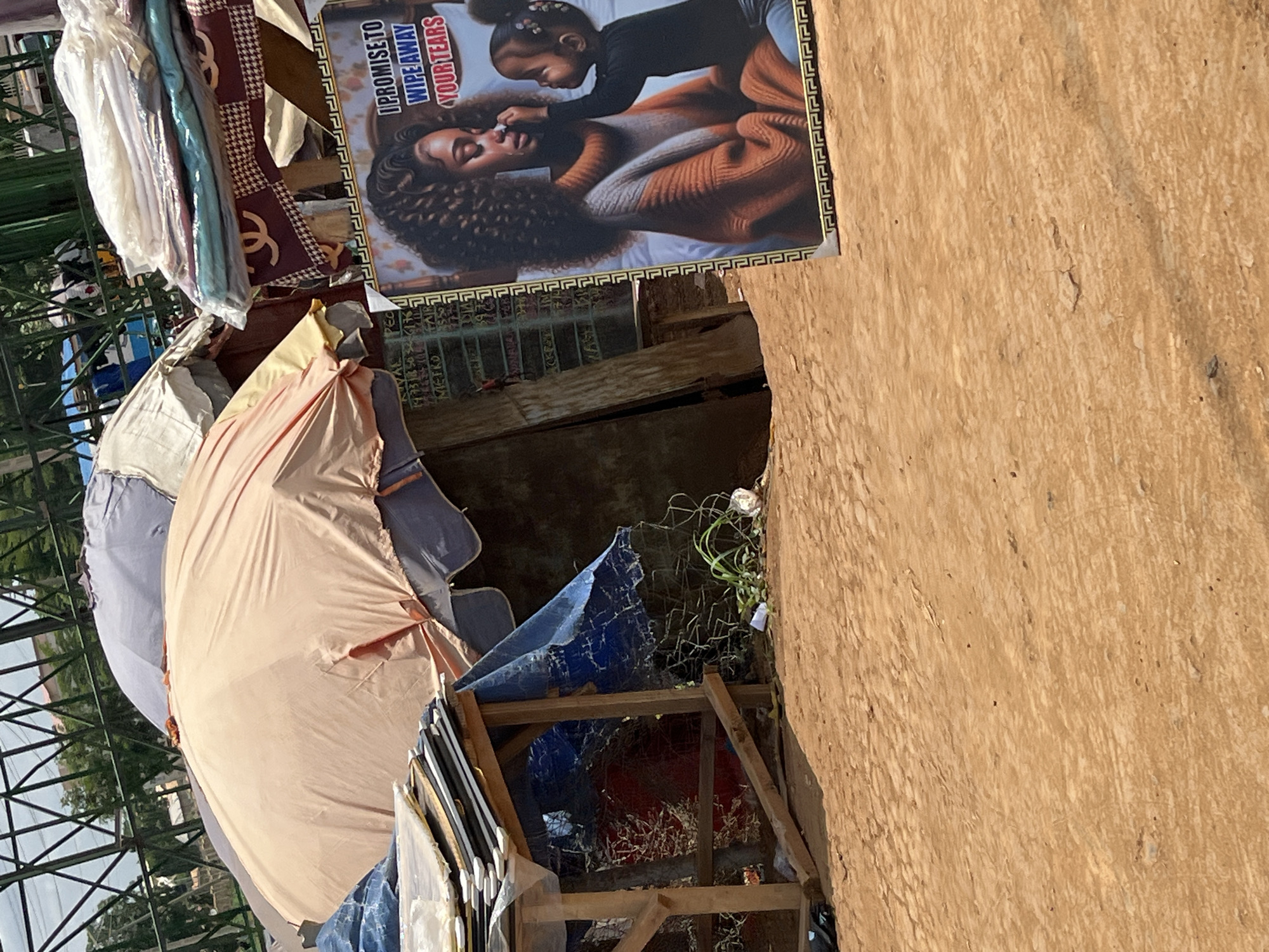 The image shows a street scene in Galadimawa Village, Nigeria. The foreground includes a stack of bedding, a pink umbrella, and what appears to be a makeshift wooden structure covered with blue tarp. A poster depicting a mother and child with the caption "I promise to wipe away your tears" is affixed to a tan-colored wall. The image depicts a sense of poverty and limited resources in the environment.