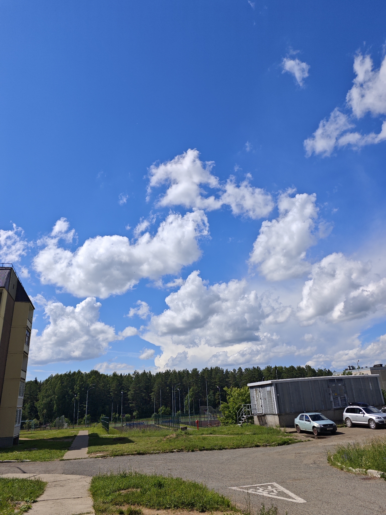 The image depicts a residential area in Markovsky, Russia, under a bright blue sky with scattered white clouds. In the foreground, there is a paved road and a sidewalk. To the left, a portion of a multi-story apartment building is visible, characterized by its light yellow facade and a dark sloped roof. Adjacent to the building, a patch of green grass and a path leading towards a playground area with sports facilities are present.
Further into the scene, a large, utilitarian gray building with a corrugated metal exterior stands on the right. Two cars are parked in front of this building: a light blue sedan and a silver SUV. Beyond the buildings and the immediate grounds, a dense forest of green trees forms a prominent backdrop. Streetlights are visible throughout the area, suggesting a well-developed neighborhood. The overall impression is one of a typical summer day in a suburban environment.