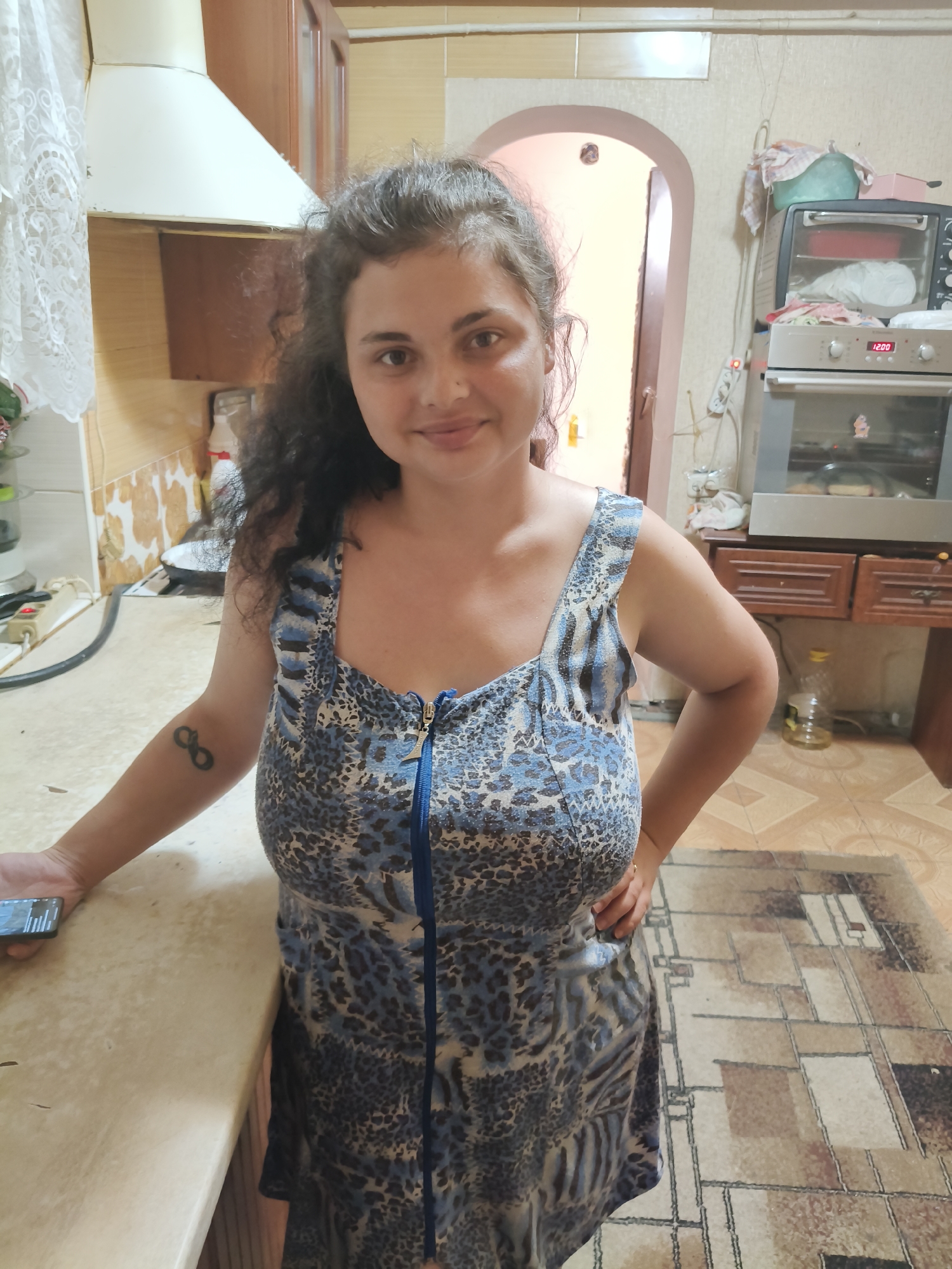 A woman with dark, curly hair is standing in a kitchen. She is wearing a blue and black leopard print dress with a zipper down the front. She has a tattoo of an infinity symbol on her left forearm. The kitchen has wooden cabinets, a tiled backsplash, and a patterned rug on the floor. There is a stove and an oven in the background, as well as a bottle of oil and a roll of paper towels. The woman is holding a smartphone in her left hand. The scene appears to be indoors in Chornobaivka, Ukraine.