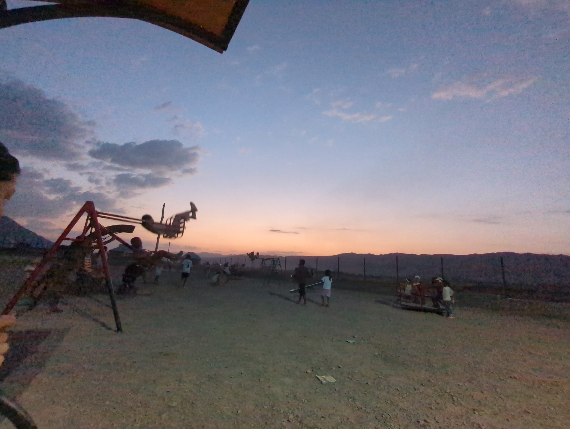 The image captures an outdoor scene in the village of Uch-Döbö, Kyrgyzstan, during twilight. A playground with a red metal swing set is visible on the left side of the frame. Several children are scattered across the dusty ground, some appearing to be playing or interacting near the swing set and another smaller playground structure on the right. The sky is a gradient of soft blue and orange hues, indicating the sun has recently set or is about to rise. Hills form the background, and a wire fence runs horizontally across the middle ground. The ground surface is predominantly dirt and gravel, with some scattered debris.