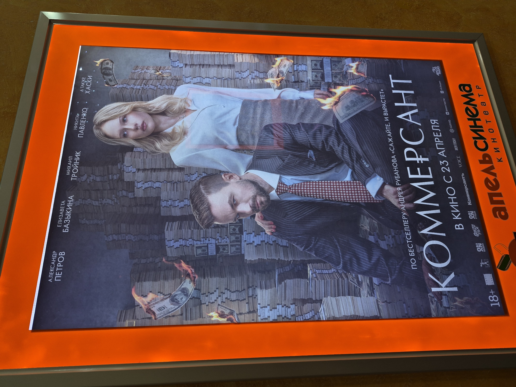 The image shows a vertically oriented, illuminated movie poster for a film titled "Коммерсант" (Kommersant, which translates to "The Businessman"), promoting its release in cinemas starting April 23rd.
The poster features two individuals: a blonde woman in a white blouse and a man with a beard wearing a pinstripe suit and a red patterned tie. They appear to be superimposed on a background of stacks of money, with burning hundred-dollar bills scattered around.
Text on the poster includes the names of actors Alexander Petrov, Elizaveta Bazykina, Mikhail Troinik, and Nikita Pavlenko, as well as the name Khaski. It states that the film is based on Andrey Rubanov's bestseller "Plant it, and it will Grow." Various logos and labels are present, including "Коммерсант," "ON GONZO FILM," "VOICE," "MEN TODAY," "кино," "вконтакте," and "VK Видео." A large red logo "Апельсинема" (Apel’sinema, a cinema chain) is on the right. A label indicating "18+" is visible in the lower-left corner.
The poster is encased in a frame with an orange illuminated border, which is framed by a silver metal frame. The environment surrounding the poster appears to be a wall. The specific location in the city of Unknown, Unknown is not determinable from the image.