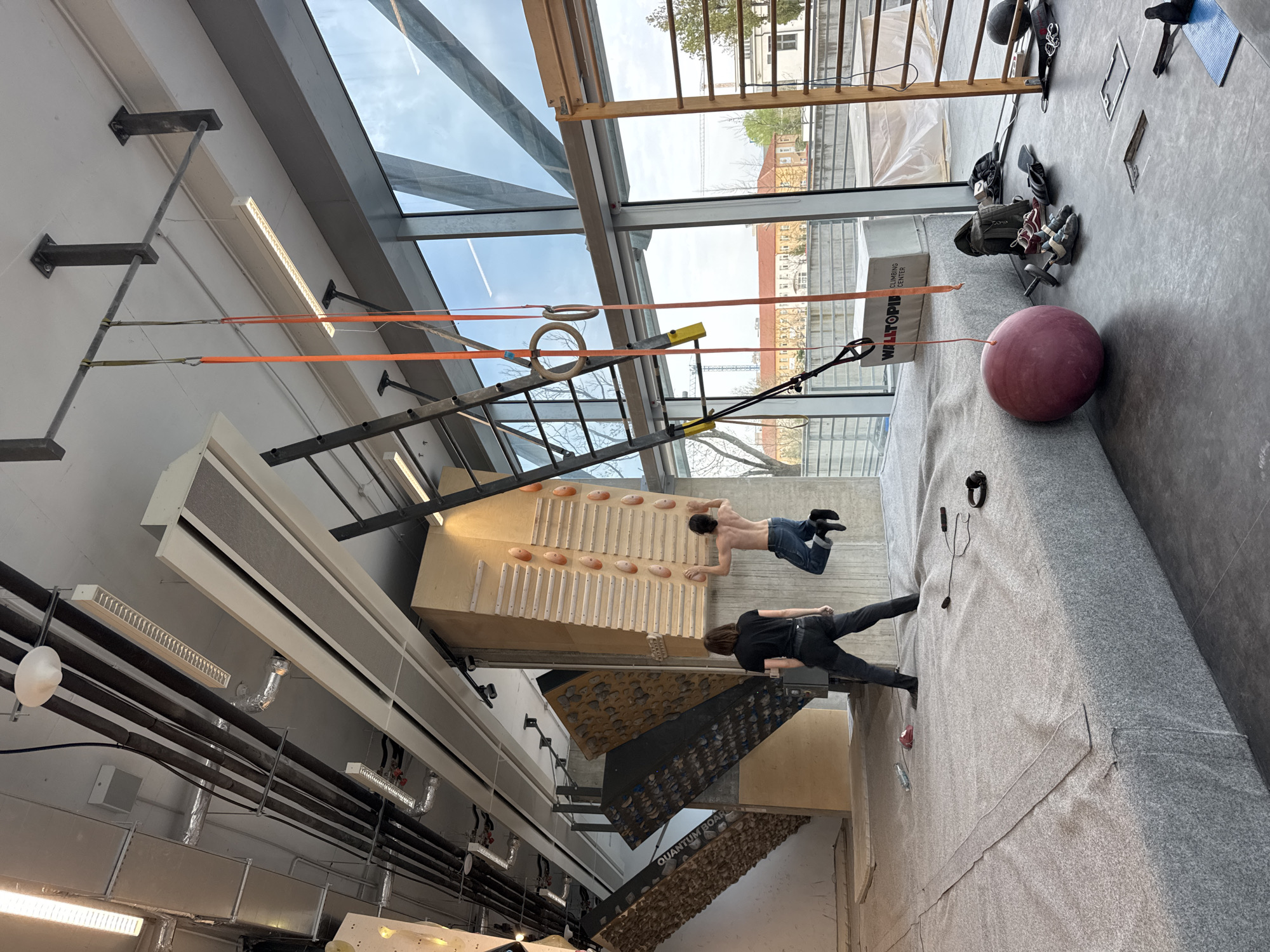 The image shows an indoor climbing gym, likely in Sofia, Bulgaria given the location of the Wаlltopia Climbing Center mentioned in the image data. The gym has a variety of climbing walls, including a wooden board with regularly spaced holds, and several other textured climbing surfaces. A man is climbing the wooden board without a shirt, wearing jeans and climbing shoes. Another person is standing on the padded floor below, appearing to spot or assist. Various training equipment is present, including gymnastic rings, a TRX suspension system, and a ladder-like apparatus. Bags, shoes, and a medicine ball are on the floor. The gym has a large skylight that allows natural light into the space. The exterior view through the skylight shows buildings and trees. There are industrial-style pipes and ventilation systems visible on the ceiling.