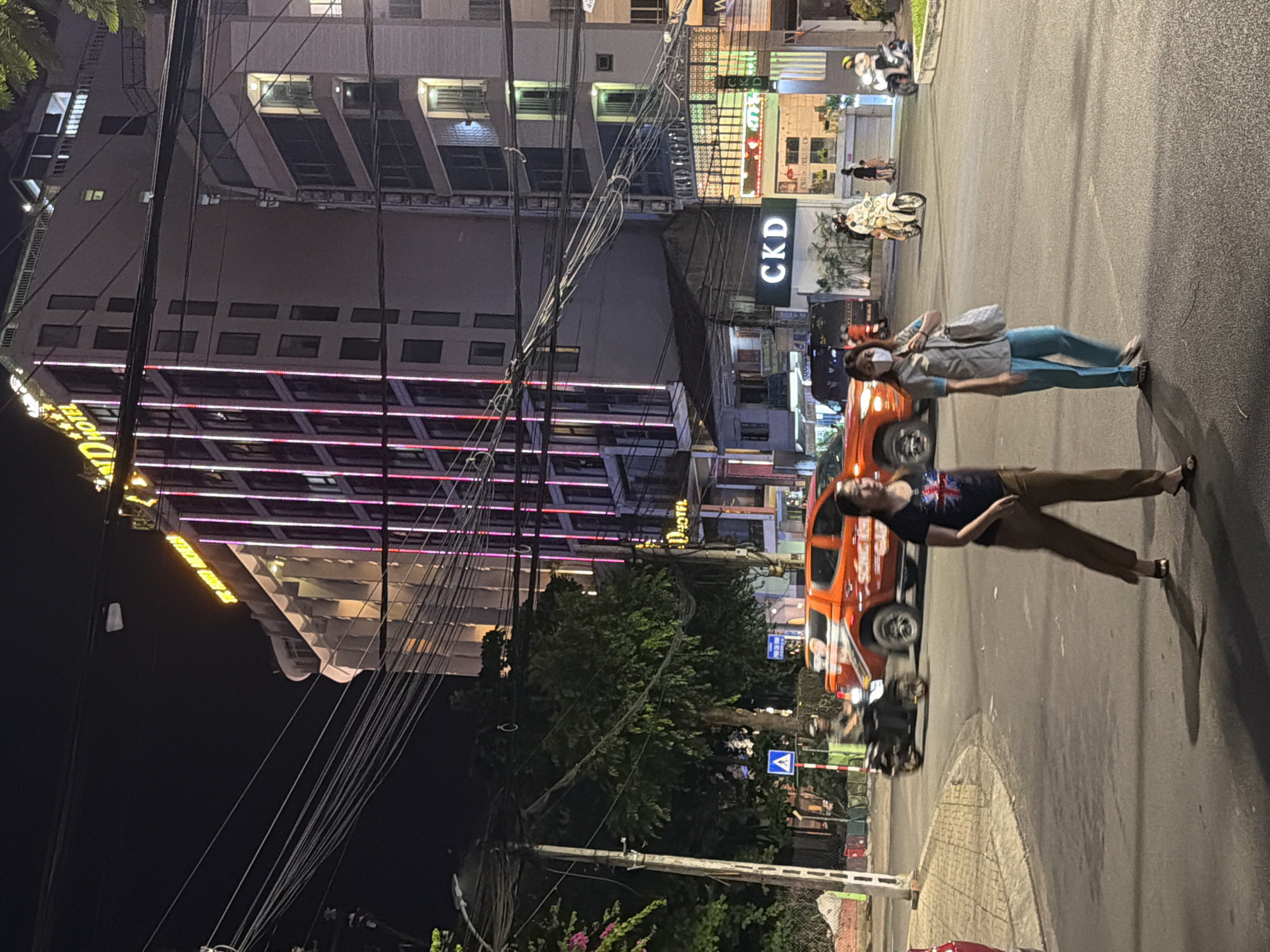 At night in Nha Trang, Vietnam, on a street corner, a woman in a black t-shirt and brown pants walks across the road while wearing a face mask. To her right, another woman in a light blue uniform and teal pants also walks across the street, carrying a bag and wearing a face mask. Behind them, an orange pickup truck drives by, and a person on a motorbike passes the truck. In the background, several buildings are illuminated with colorful lights, including a tall hotel with neon pink and white lights. One of the buildings has a sign that reads "CKD." The scene takes place on a paved road with streetlights casting shadows.