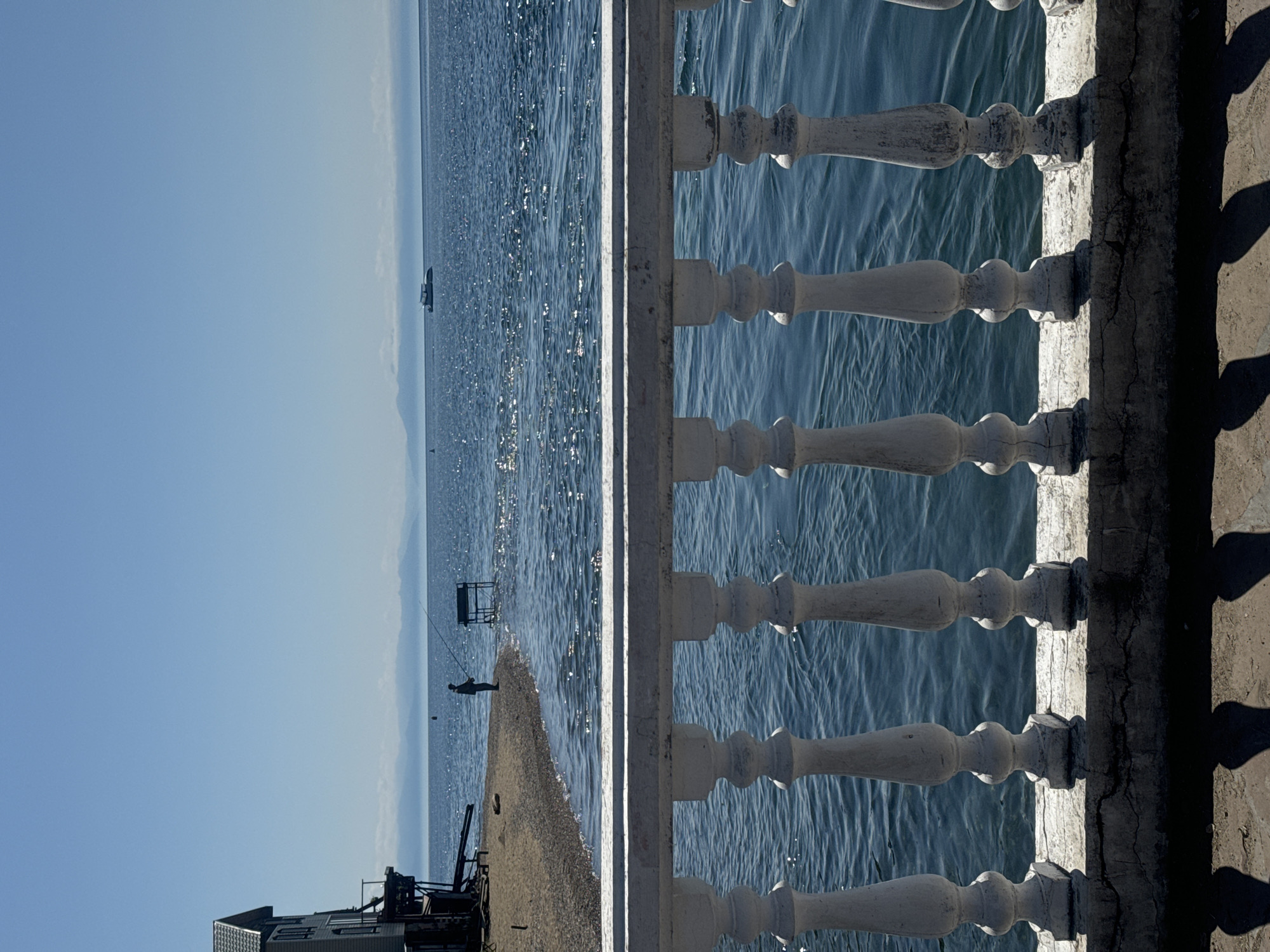 The image depicts a bright, sunny day scene overlooking a body of water, likely Lake Baikal, given the reference to Listvyanka, Russia. In the foreground, a white balustrade with ornate pillars lines the view. Beyond the balustrade, a sandy and pebbly shore leads to the water's edge. A single person is visible standing on the shore, holding a fishing rod and fishing. To the left of the fisherman, a tall, light-colored building with multiple levels and balconies stands partially on the shore. In the middle distance, a small boat with a white cabin sails on the shimmering, blue water. The horizon is marked by a hazy, mountainous landscape under a clear blue sky. The scene conveys a sense of calm and leisure, with the bright sunlight reflecting off the water and casting shadows from the balustrade onto the foreground ground.