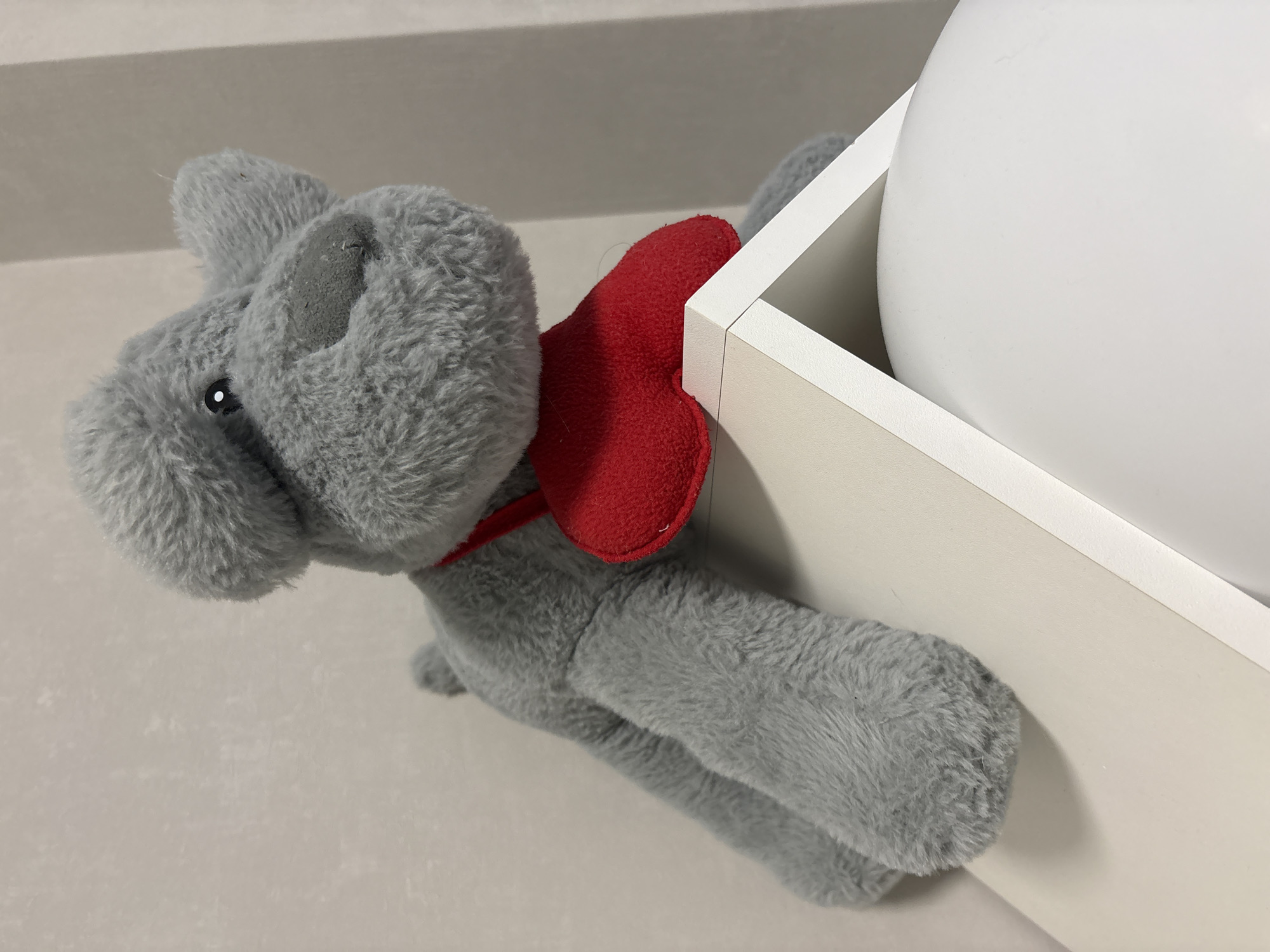 A gray stuffed animal dog with a red cape is positioned next to a white container holding a white sphere. The location is not discernible from the image.
