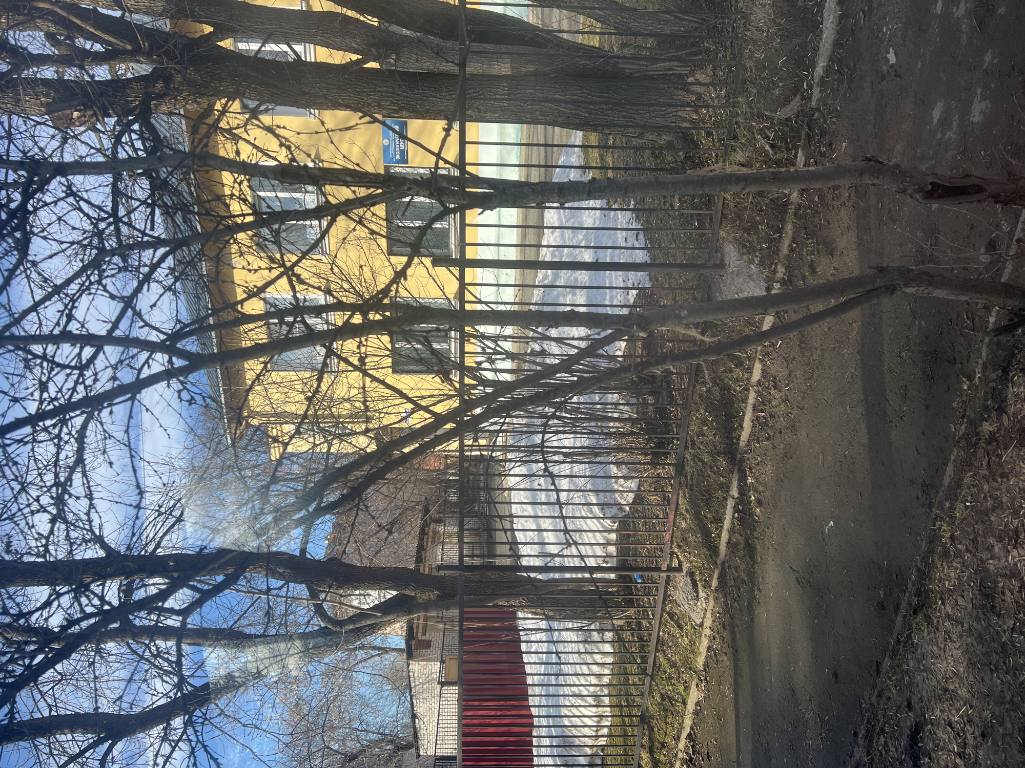 A street scene in Berezniki, Russia. A yellow building is partially visible behind bare tree branches. A black metal fence separates the building from a snowy area and a dirt path. Patches of snow are visible on the ground. Part of a red and white building is visible behind the fence. The sky is blue.