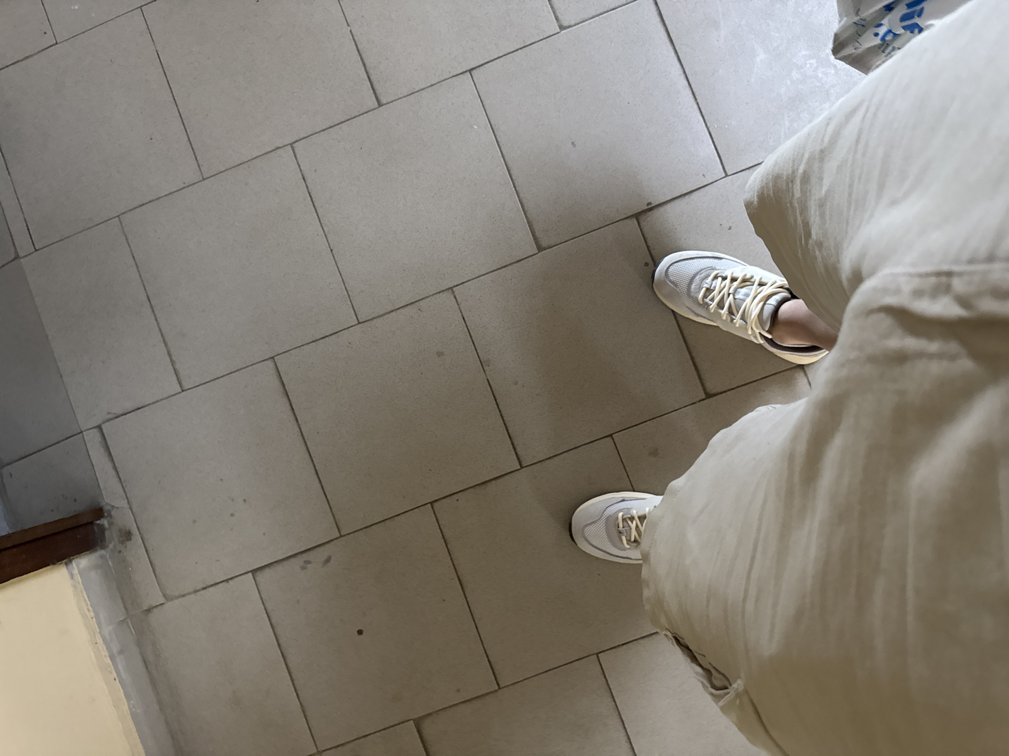 The image shows a person's legs and feet from a first-person perspective, standing on a tiled floor. The person is wearing light-colored pants and athletic shoes. The floor is composed of large, rectangular, light gray tiles with visible grout lines. The lighting suggests an indoor environment, possibly a hallway or a room with a doorway visible in the upper left corner. There are no discernible elements that would allow for identification of a specific location within the city of Sofia, Bulgaria.