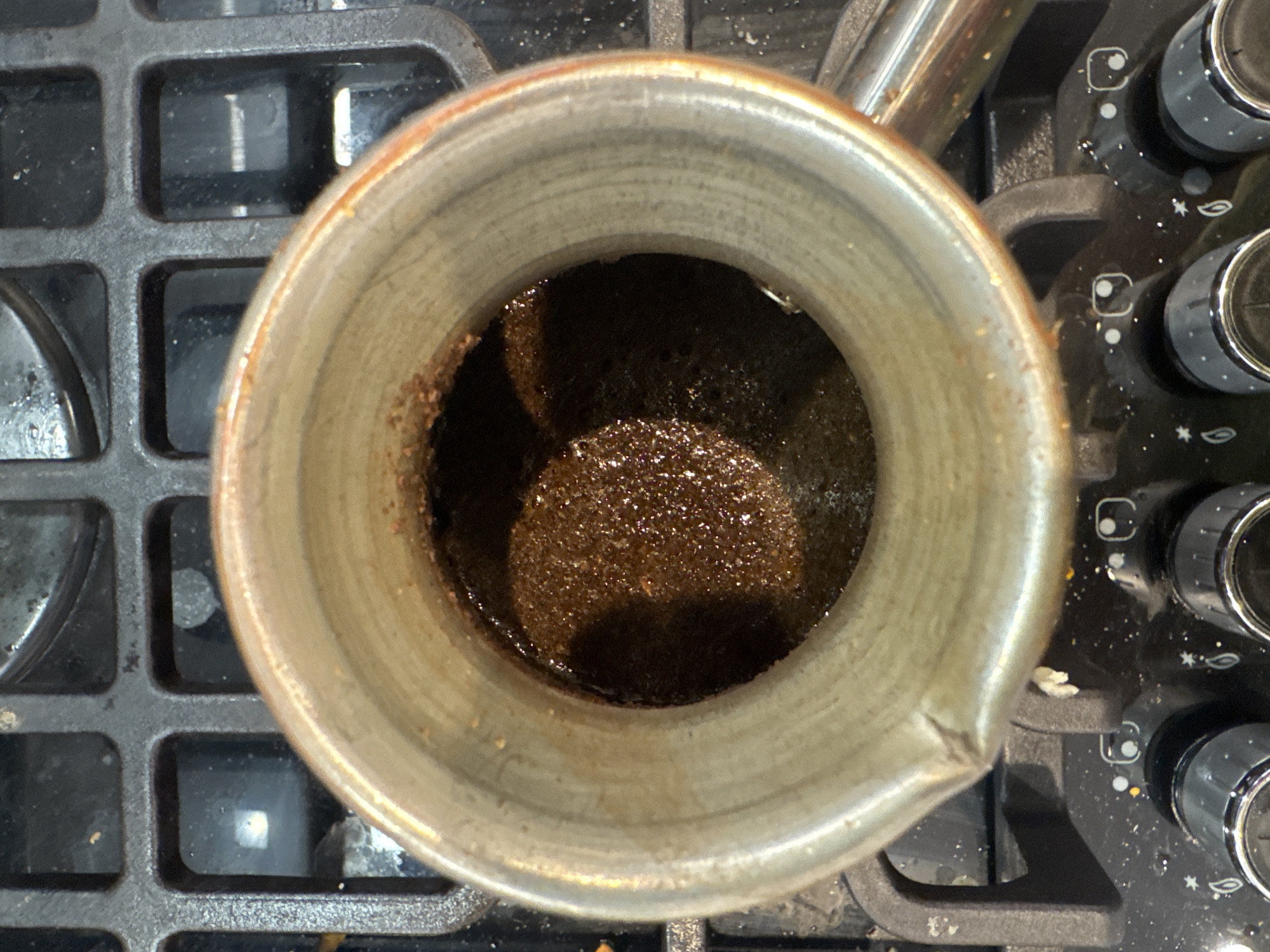 An overhead shot captures a metal Cezve, or Turkish coffee pot, filled with dark brown coffee grounds and liquid, resting on the grate of a black gas stove. The Cezve's interior shows signs of use, with a slightly tarnished or stained appearance on its metallic walls, and the coffee mixture has a fine, bubbly texture.
The stove's black surface features a sturdy, dark-colored metal grate with a square grid pattern, supporting the Cezve. To the right, two control knobs are partially visible. These knobs are black with shiny silver tops and display white symbols: a circle with a dot, indicating a burner, and small dots around it, likely denoting heat levels. Another symbol, resembling a flame or leaf, is also present near one of the knobs. The stove surface itself has scattered white specks, possibly dust or residue.
The scene suggests the initial stage of preparing Turkish coffee in a kitchen setting. No people are visible, and no specific time of day or emotions are conveyed. No discernible text is present, only symbolic markings on the stove controls.
