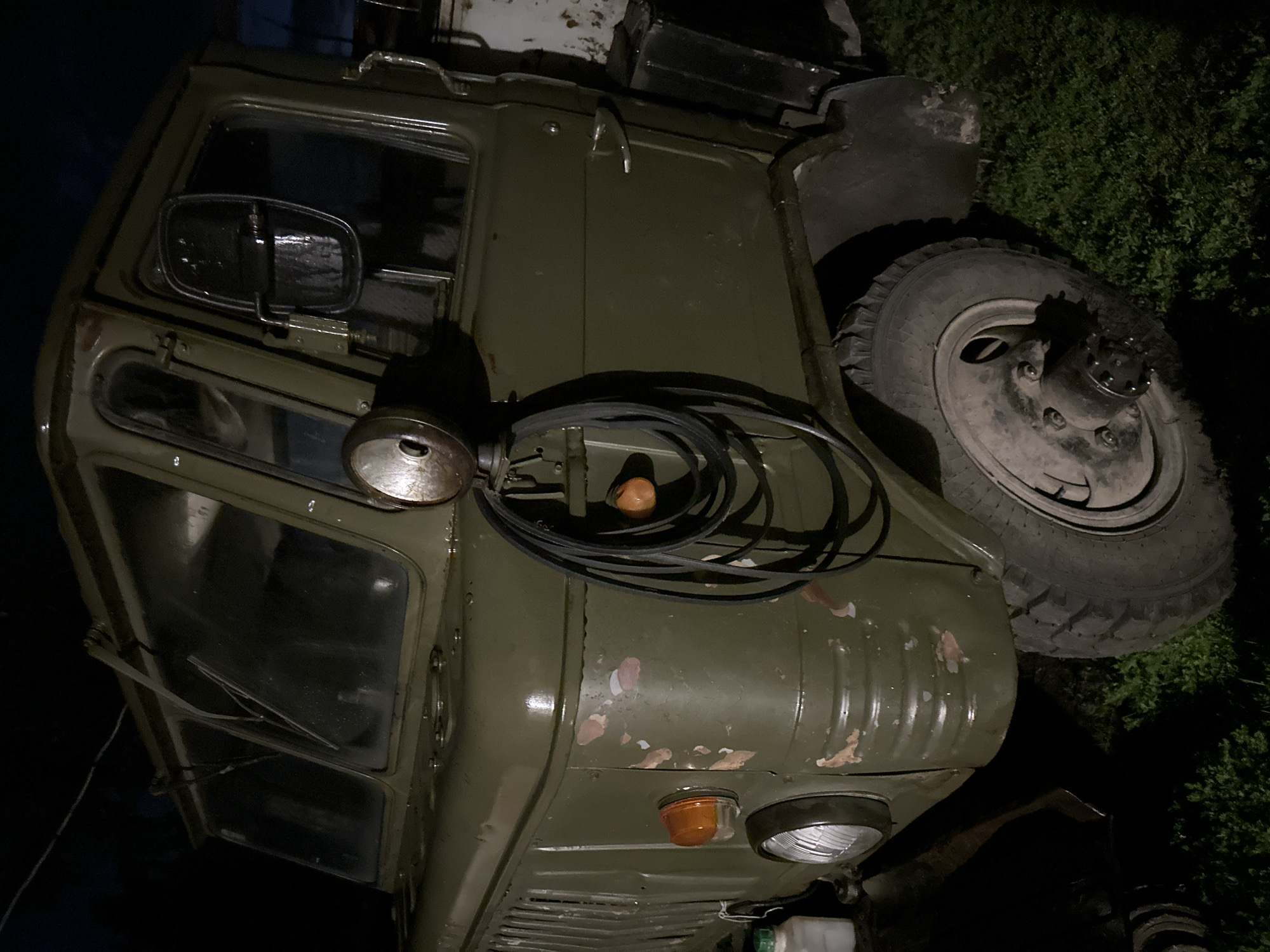 An old, olive-drab military truck is parked on a grassy area at night. The truck has a heavy-duty build with large tires and a rugged exterior. A spare tire, coiled and attached to the side of the truck, is visible. The truck's headlights and an orange turn signal are illuminated. The overall impression is one of a weathered, utilitarian vehicle in a natural, outdoor setting.