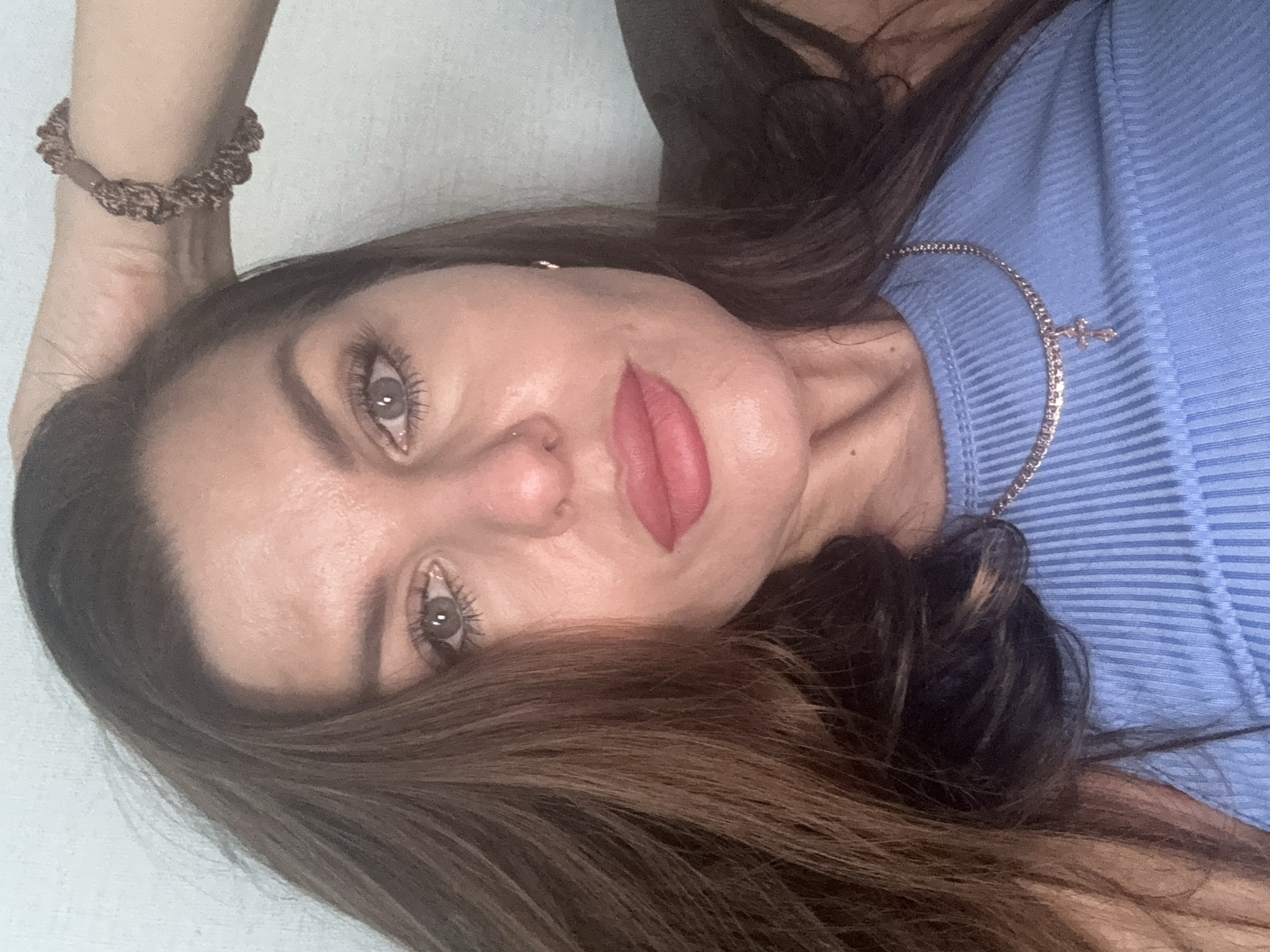 A woman with brown hair, fair skin, and grey eyes is visible. She is wearing a blue ribbed top and a delicate necklace with a cross pendant. Her lips are painted with red lipstick. A brown scrunchie is on her wrist. The image is taken at an oblique angle. The location in Chaikovsky, Russia, is not discernible from the image.