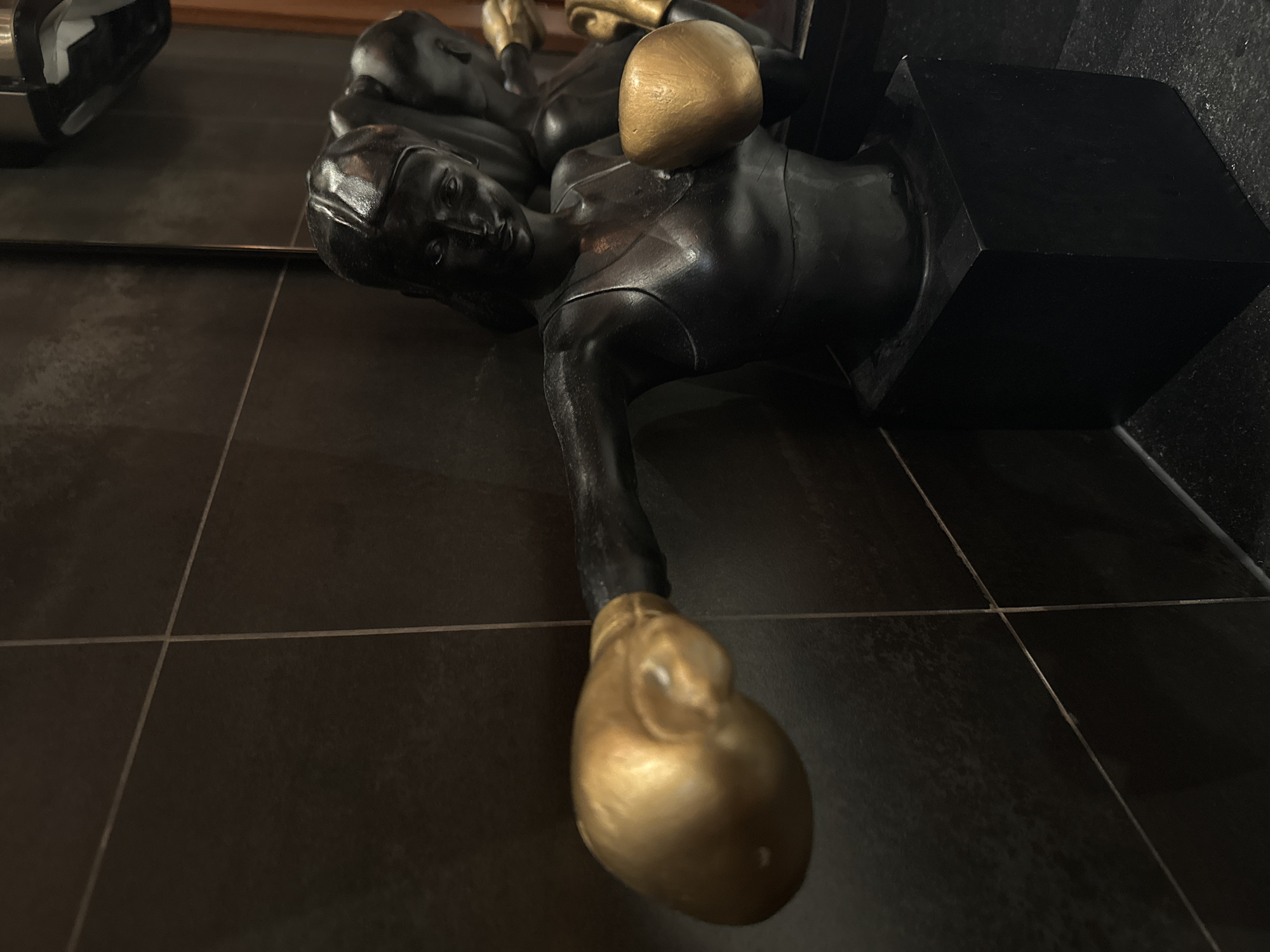 A black statue of a female boxer stands on a black pedestal. The boxer is depicted from the torso up, wearing a sports bra and boxing gloves. The gloves are gold, and the boxer's skin is black. Her left arm is extended forward as if throwing a punch, and her right arm is bent, with her fist held near her chest. Her head is turned slightly to the left. Behind the main statue, another partial statue of a female boxer is visible, also wearing gold boxing gloves. The statues are positioned against a dark, tiled wall, which appears to be in a bathroom or similar indoor setting. The lighting is dim, casting shadows on the figures and the wall.