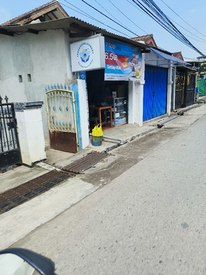 The image shows a small storefront in Padalarang, Indonesia, located at latitude -6.851 and longitude 107.474. The storefront has a blue painted facade with a large sign that says “Fresh Zone Laundry” with a laundry machine logo and a price list for washing clothes. There is a blue door to the right of the sign, as well as another door behind the sign that appears to lead to a back room. A yellow plastic bin is sitting next to the door on the right, with a dark grey pipe running along the wall. There is a drain in the middle of the sidewalk in front of the storefront, with a concrete road in front of it. There is a small, white, residential building to the left of the storefront, with a metal gate leading to a small yard. It appears to be a bright, sunny day with power lines running along the street.